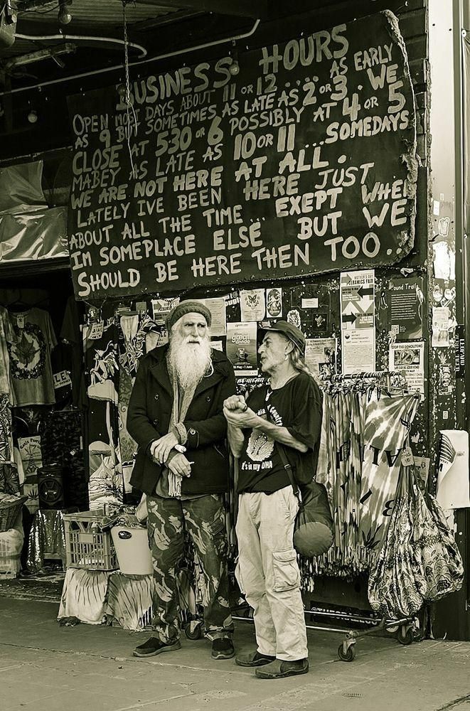 This very old black and white photo captures a street scene featuring two individuals standing in front of a shop with a unique, hand-painted sign detailing its "Business Hours."

The sign humorously describes erratic opening and closing times, stating, "OPEN MOST DAYS ABOUT 11 OR 12 AS EARLY AS 9 BUT SOMETIMES AS LATE AS 2 OR 3. WE CLOSE AT 5:30 OR 6 POSSIBLY AT 4 OR 5 MABEY AS LATE AS 10 OR 11... SOMEDAYS WE ARE NOT HERE AT ALL."

It further adds, "LATELY IVE BEEN HERE JUST ABOUT ALL THE TIME EXEPT WHEN IM SOMEPLACE ELSE BUT WE SHOULD BE HERE THEN TOO."

The two individuals appear to be conversing, with one sporting a long white beard and the other wearing a cap and holding something