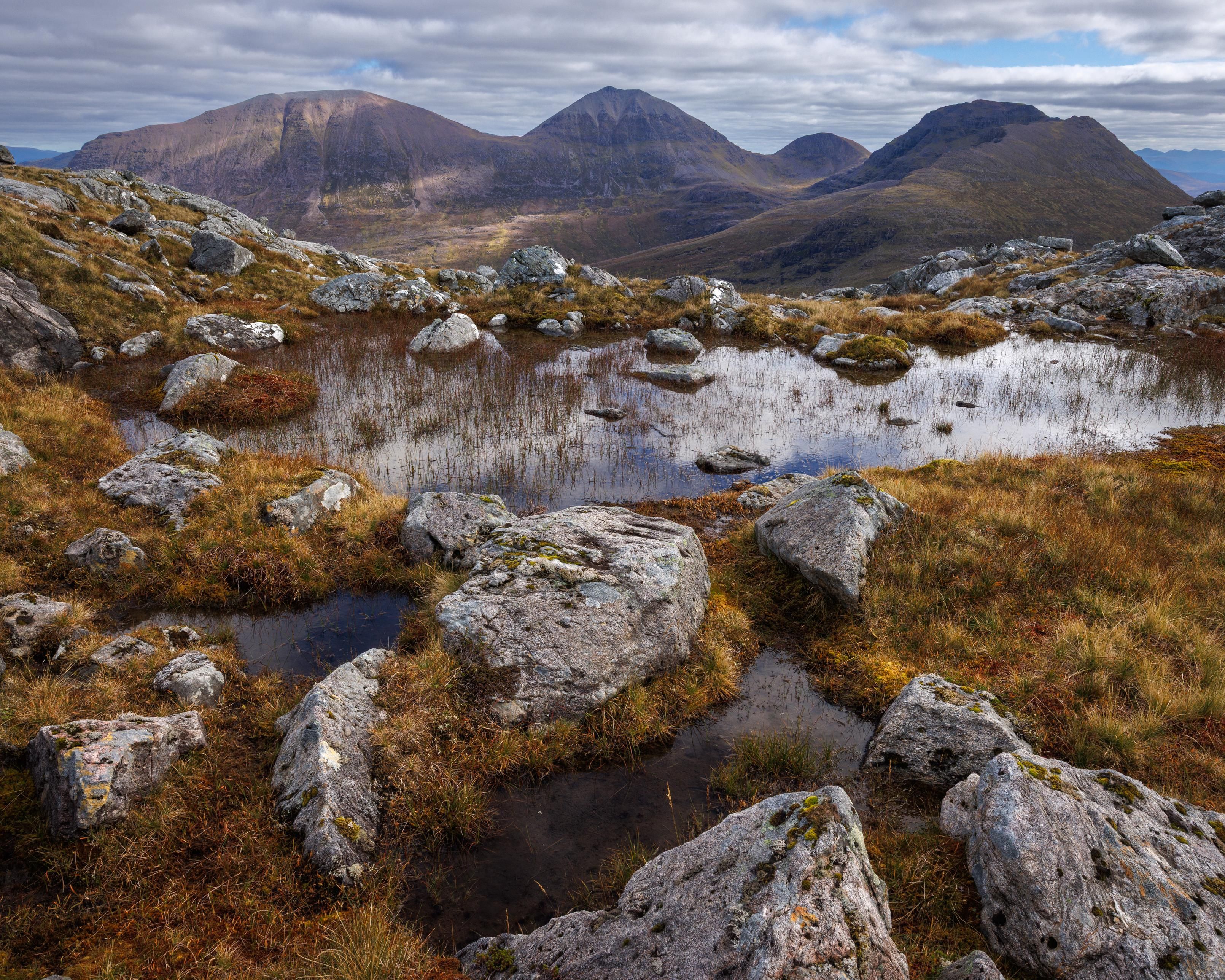 a boggy moorland of grasses and rocks dominate the foreground of this mountain image taken in remote scotland. white gneiss boulders interact with golden grass and reeds. the small lochan is perfectly still and reflects the partly cloudy skies above. three distant mountains stand tall in the background with a prominent central peak. the whole scene is dotted in a patchwork of light
