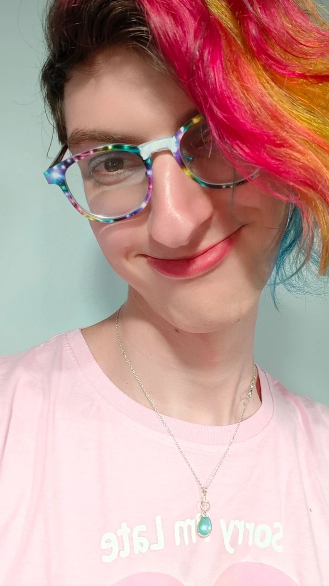 A white girl with pink, yellow, and blue hair.  Her glasses are covered in stars, and she's wearing a raindrop necklace and a pink shirt.