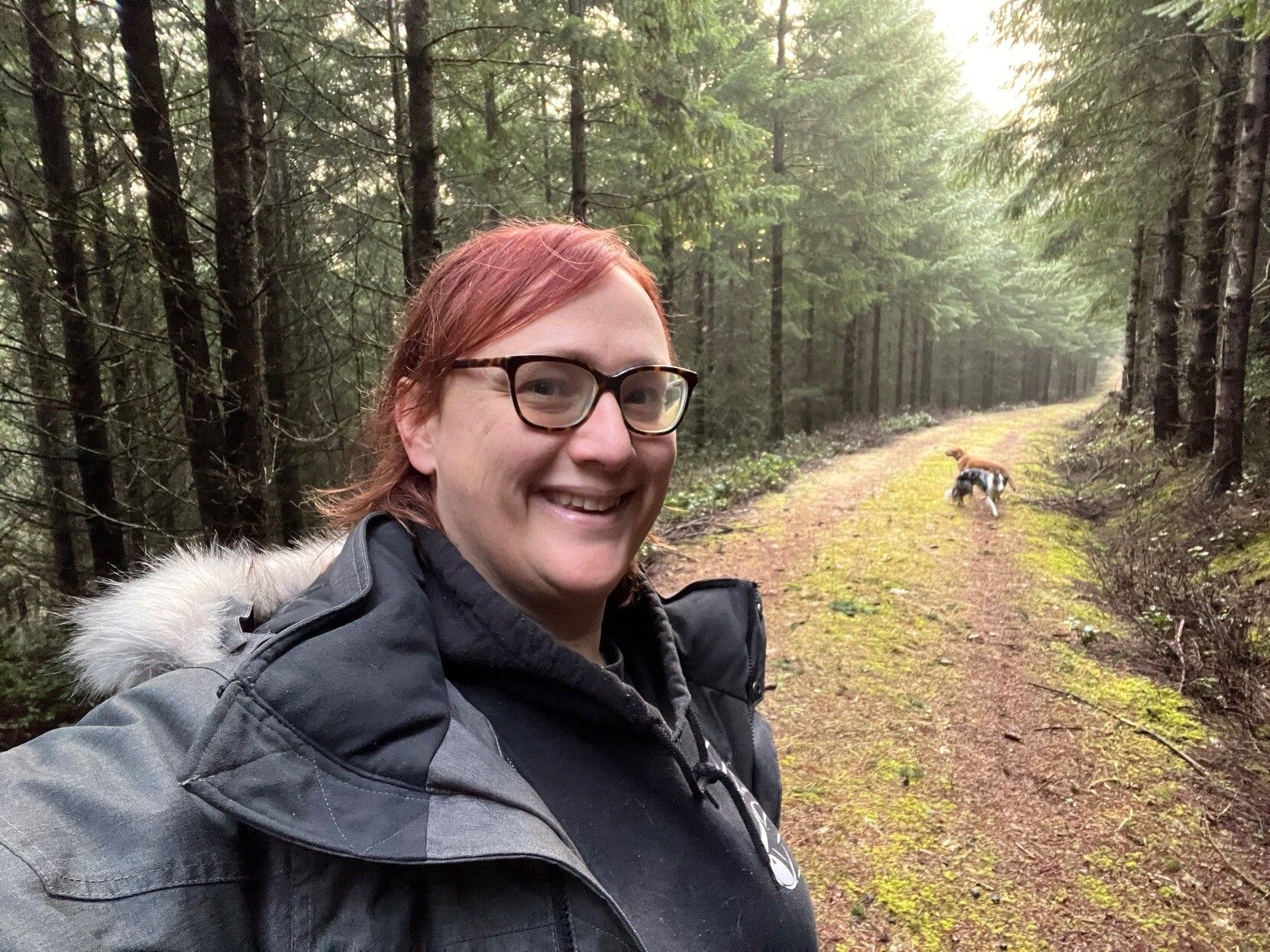 Nikki (@Aminorjourney) with her dogs on an early winter walk. She is wearing a thick winter coat with a fuzzy hood, has red hair brushed back, and thick-rimmed glasses. She is on a long forestry trail with tall trees on either side, while  a border collie and Labrador play in the distance. 