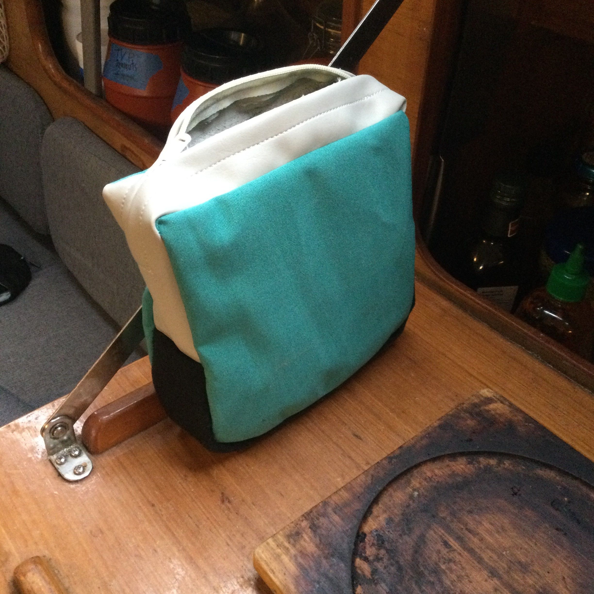 a little square bag, with a thinner edge and a zipper closing overtop. The front and back are teal, the bottom thickness of the bag is black, and zipper plaque is light grey.
The bag is sitting over a wooden pantry door, with food visible in the background.