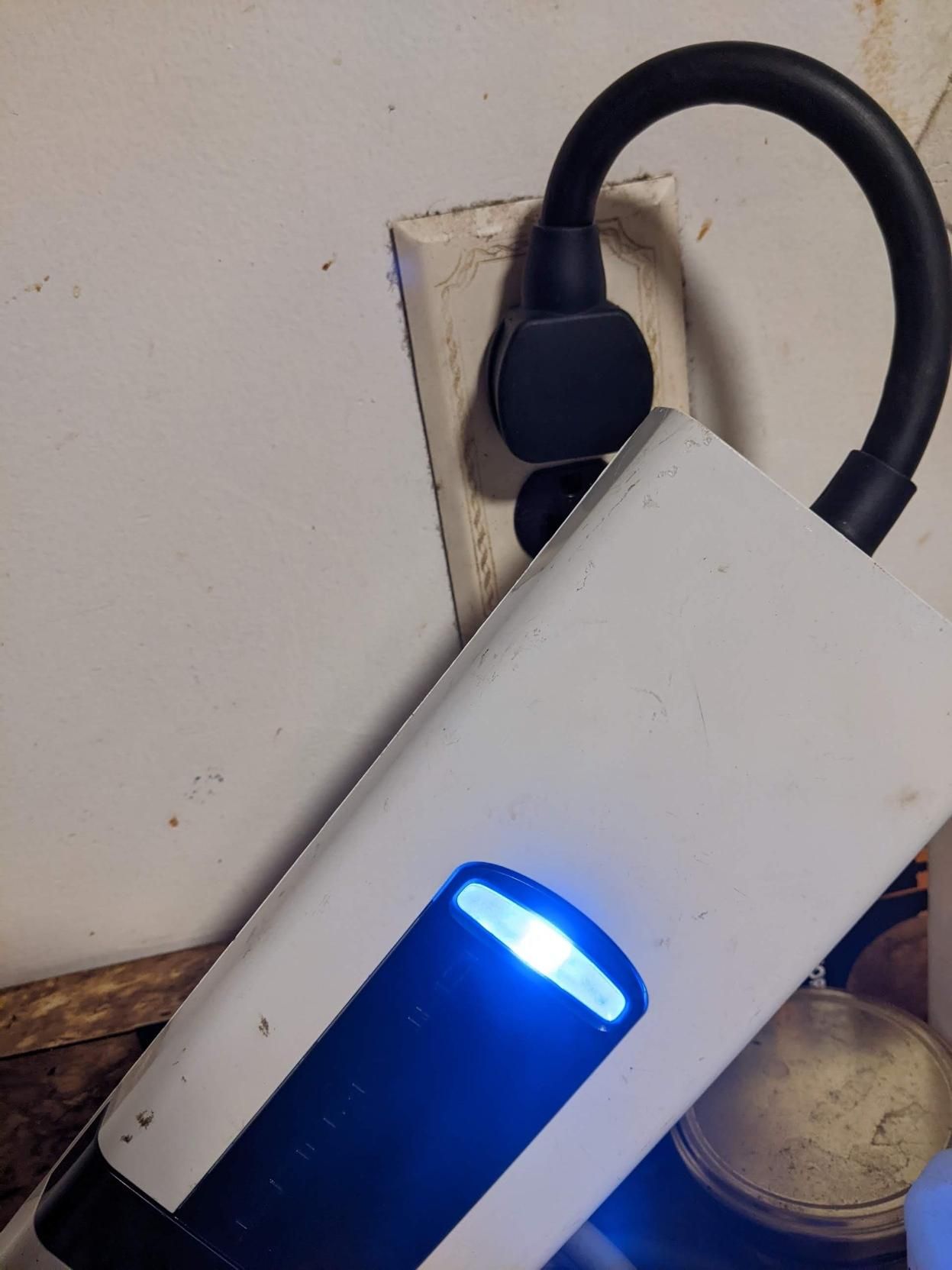 Big white rectangular object with blue light in the middle and one end of the pack with a thick black wire bent and plugged into a wall socket