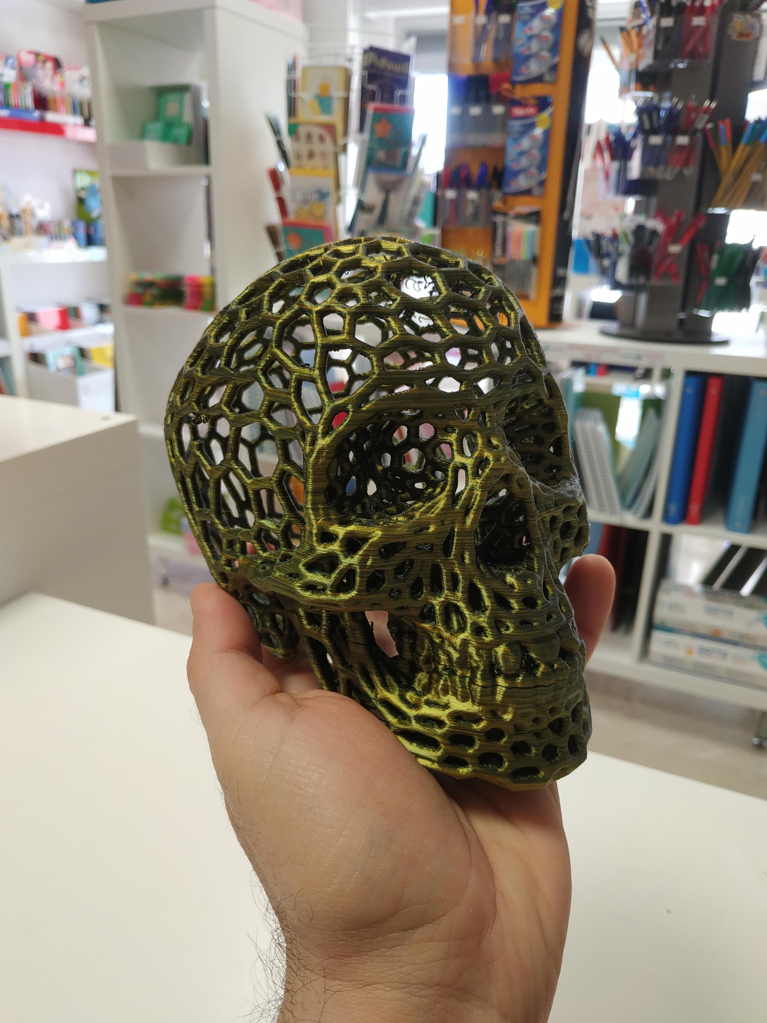 Foto di una mano (la mia) che sorregge un teschio stampato in 3D con un filamento giallo e oro. Il teschio ha una struttura reticolare e non piena. Sullo sfondo una cartoleria.