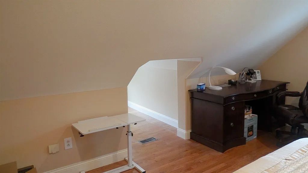 An attic room with a steeply sloped ceiling and a small opening next to a desk. It's very small and has a heating duct in the floor right in the middle of the opening. 