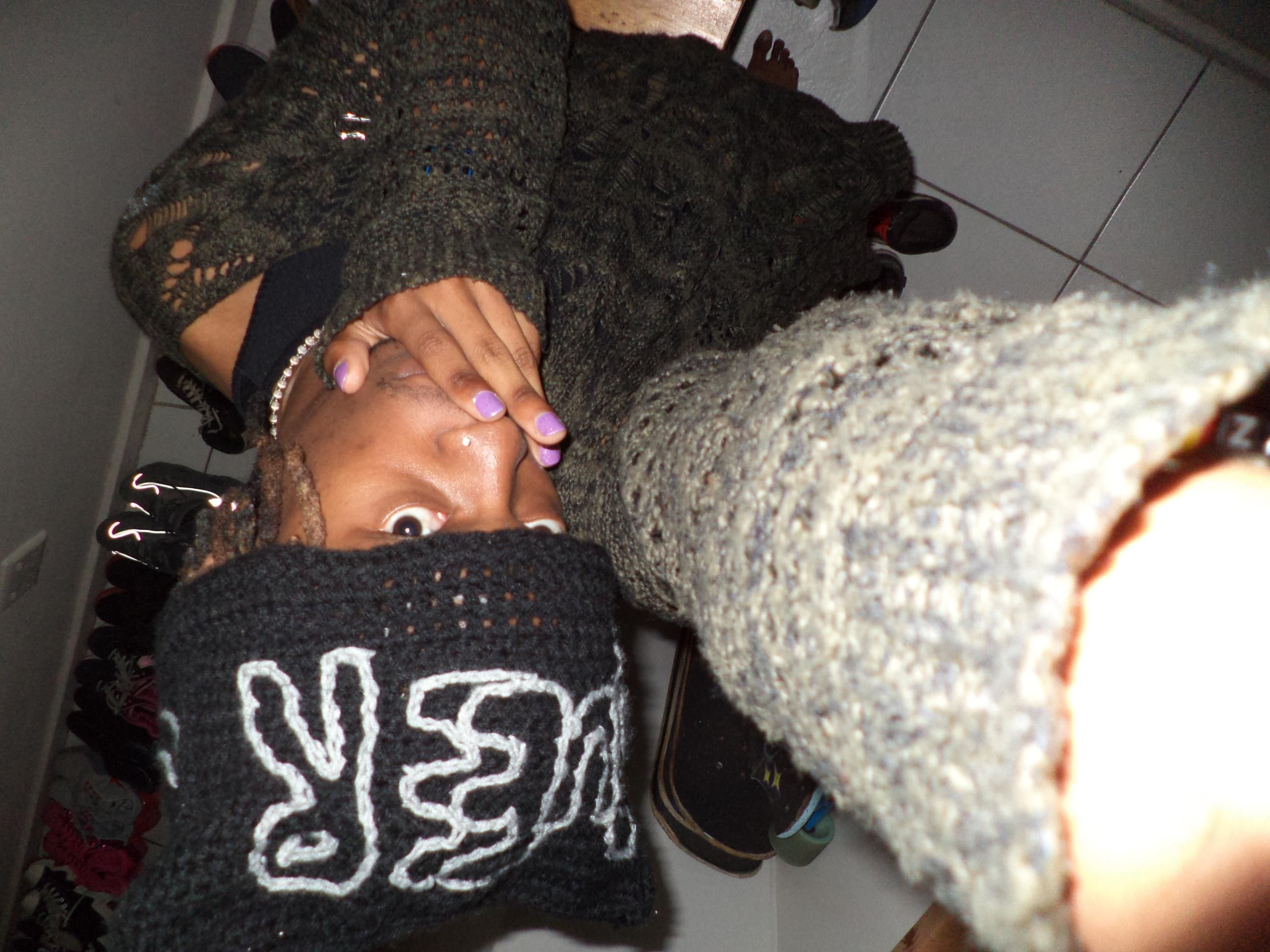 a cute selfie of me sitting with a green knit top and black skirt, with a cute crochet hat with the words "queer". And purple nail. The camera is held above my head pointing down to me.