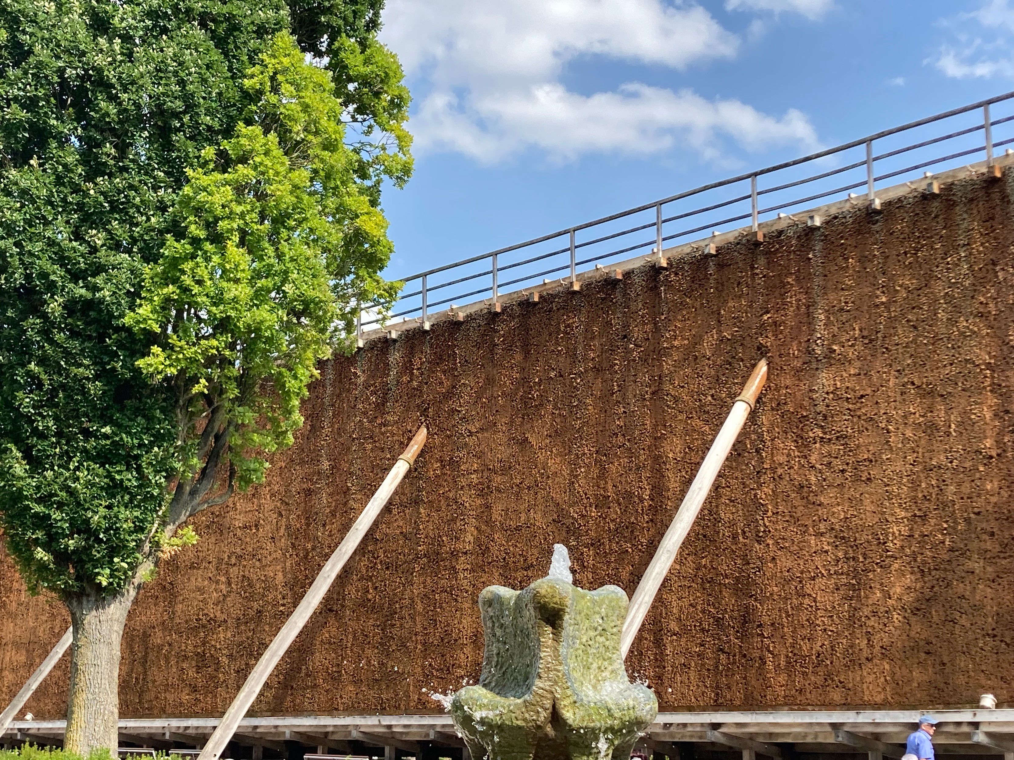 Teil eines Gradierwerks, sehr hoch, Wasser tropf langsam herunter. Im Vordergrund die Spitze eines sprudelnden Wasserbrunnens.