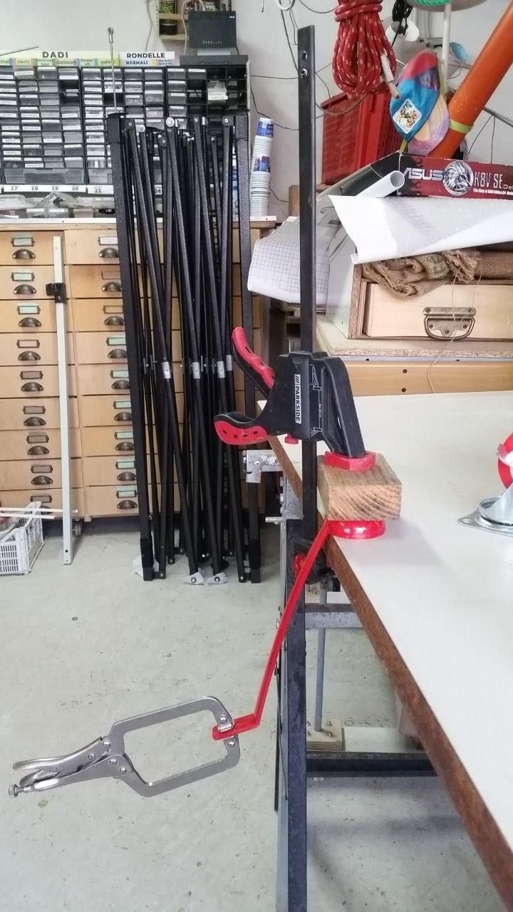 Mestolo di plastica rossa morsettato al bordo di un tavolo.
Al fondo del manico, che senza peso è inclinato a 30°, c'è una pinza a scatto per saldatura.