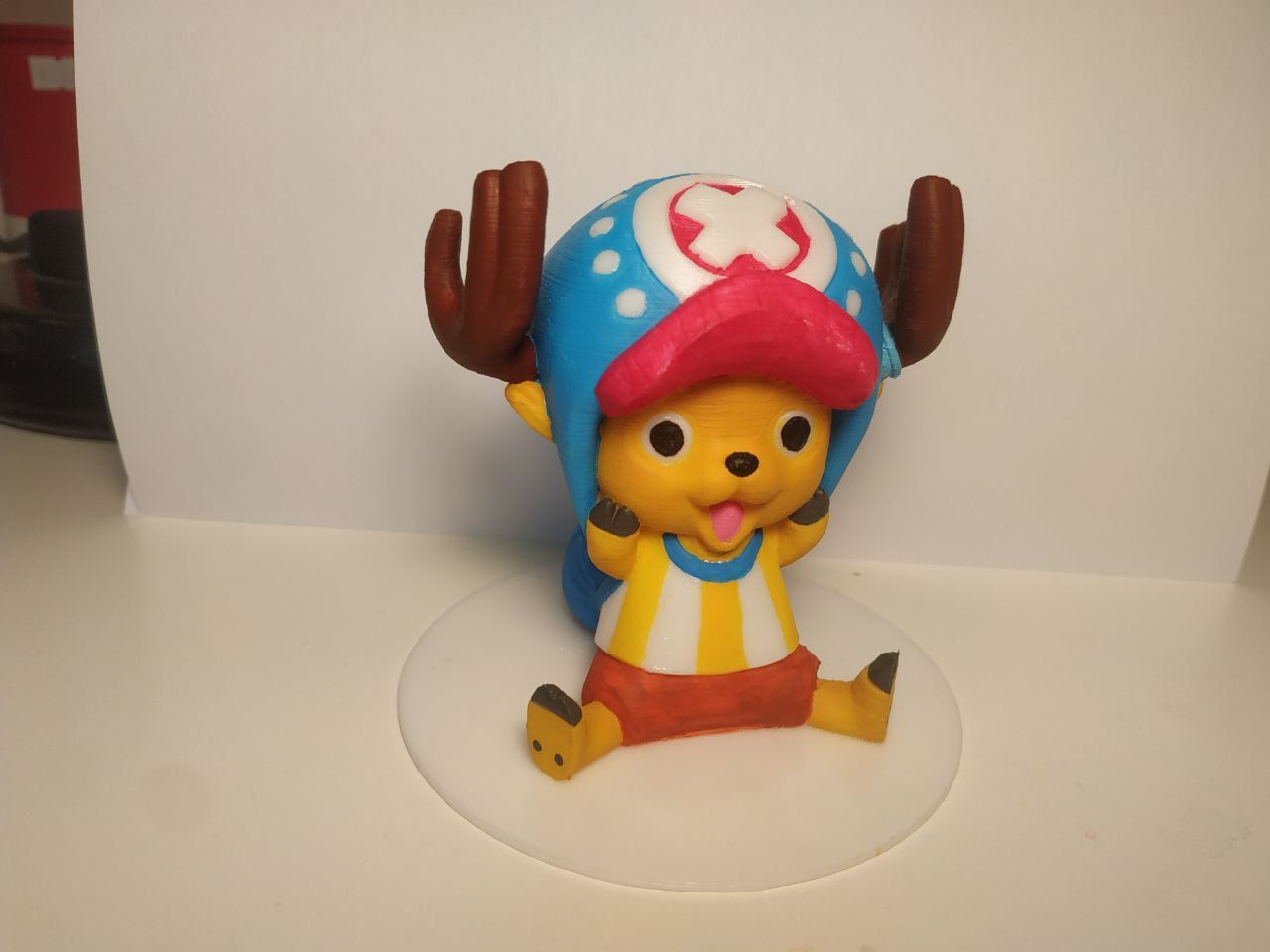 Modellino stampato in 3d del personaggio di One Piece Tony Chopper.
Seduto su una base tonda e bianca, con le zampine in faccia e espressione sorpresa e felice. Cappellone azzurro con croce bianca su sfondo rosso e con visiera rossa, magliettina a righe gialle e bianche. Corna marroni.
