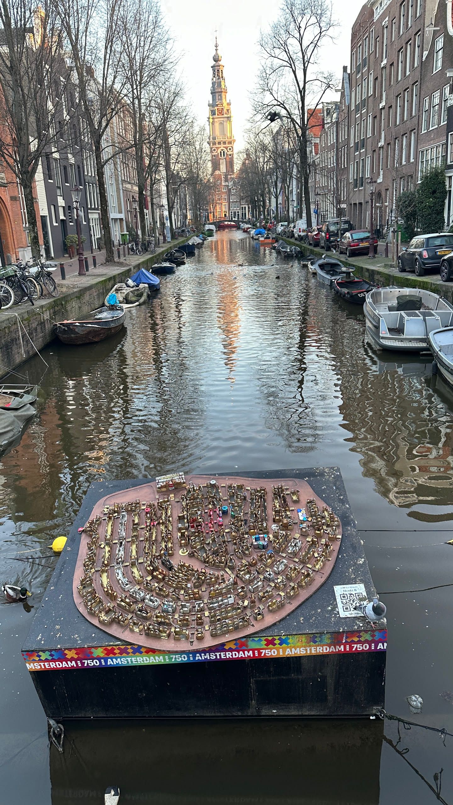 black cube floating in a canal with hundreds of small locks arranged to look like the rings of amsterdam. zuiderkerk is in the background, reflectd in the water