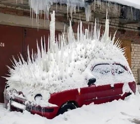 A red car parked outdoors in winter is completely covered in snow and long, sharp ice spikes, forming a dramatic, porcupine-like silhouette. Thick icicles hang from the roof and hood, while the windshield and mirrors are frozen over, suggesting prolonged exposure to extreme cold. Snow blankets the ground around the vehicle, and the car is parked in front of an industrial-style building with brick walls and a large metal door, reinforcing the harsh, icy conditions.