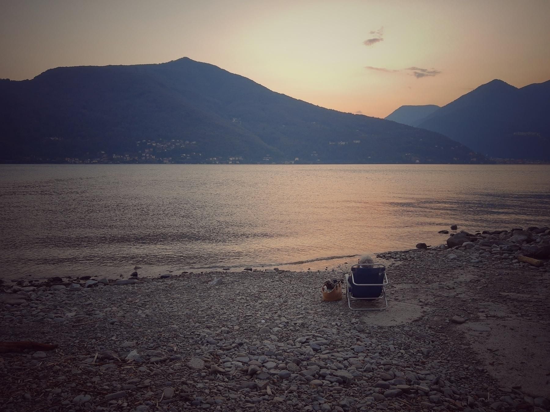 Una persona siede su una spiaggia di ciottoli al tramonto, di fronte a un lago tra le montagne.