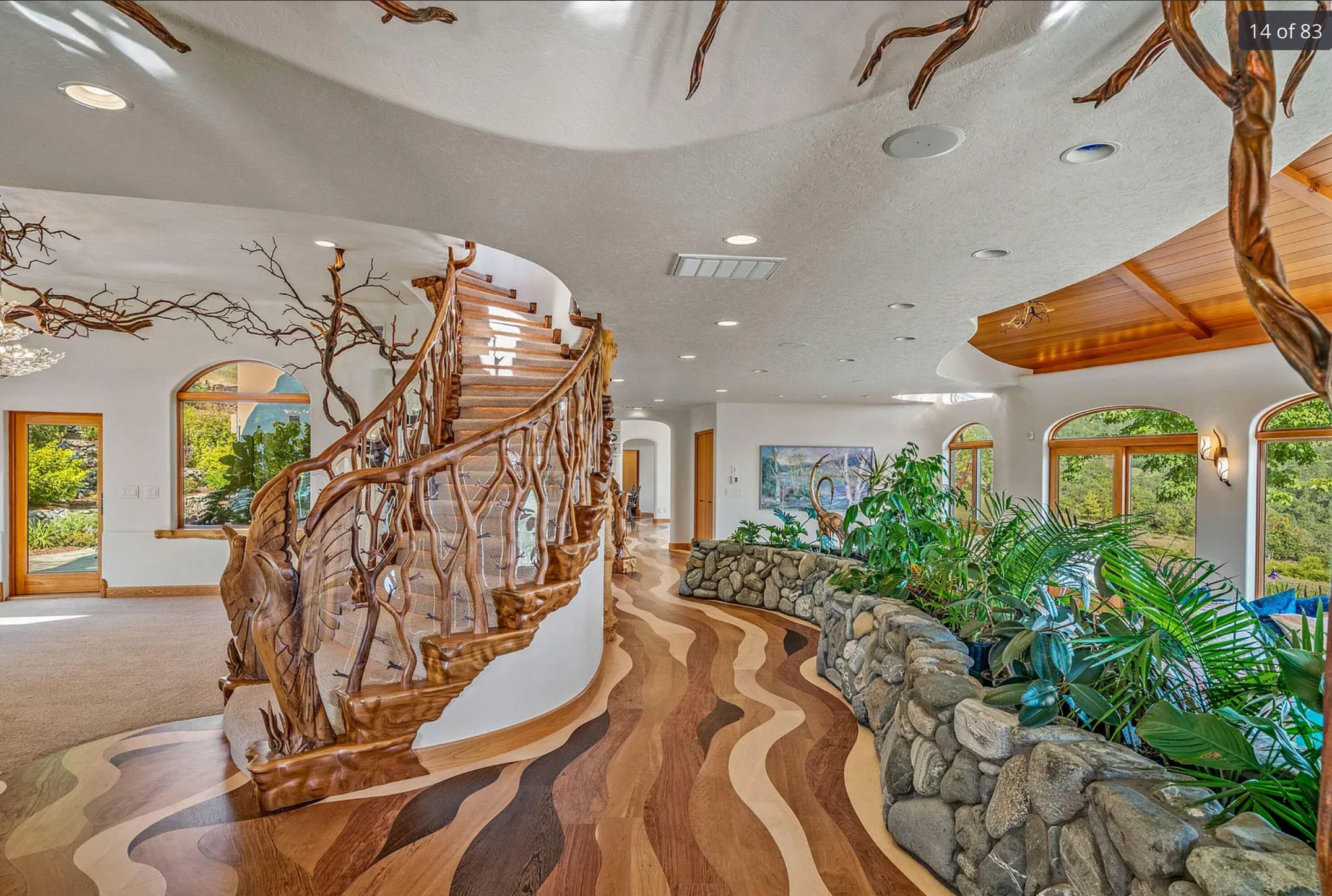 A view of the inside showing a wood floor with curved and winding stripes, almost like a river. There is a stone retaining wall with green plants inside it, and an ornate stairway made of branches. There are also branches across the ceiling in the distance. 
