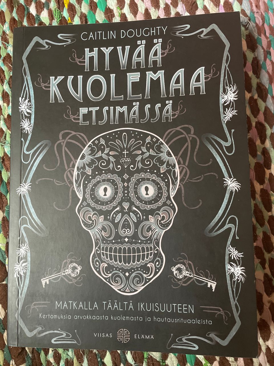 Tumma kantinen pokkari. Keskellä koristeellinen Días de los Muertos-tyylinen piirros pääkallosta. silmäkuopissa avaimen reiät. kukkia ja muita kuvioita kuin tatuointeja.