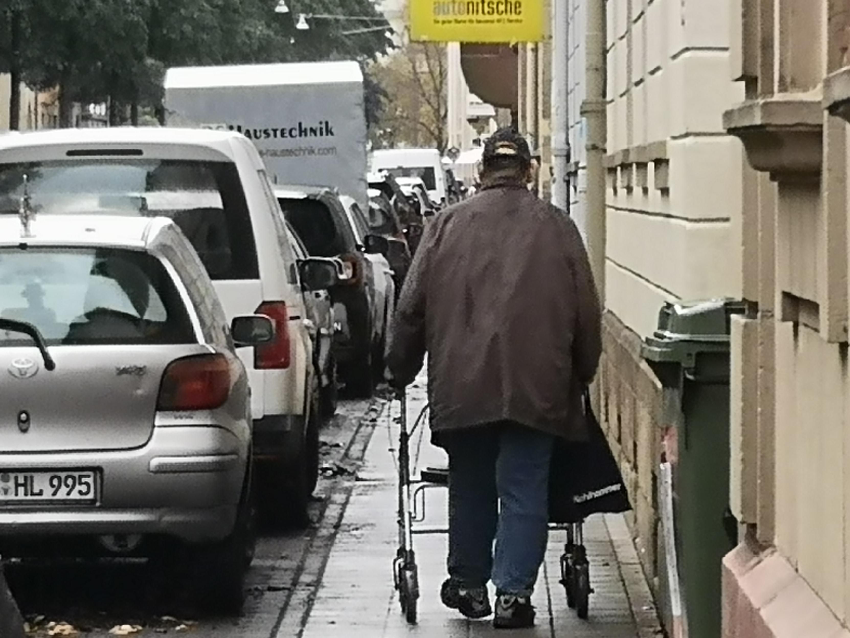 Eine ca 1 Meter breite Gehwegschlucht, zwischen geparkten Autos und Häuserwand schiebt eine Person einen Rollator 