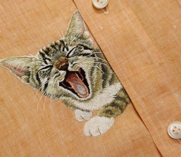 Realistic embroidery of the upper half of a tabby cat with moujth open and eyes closed, on an orange shirt