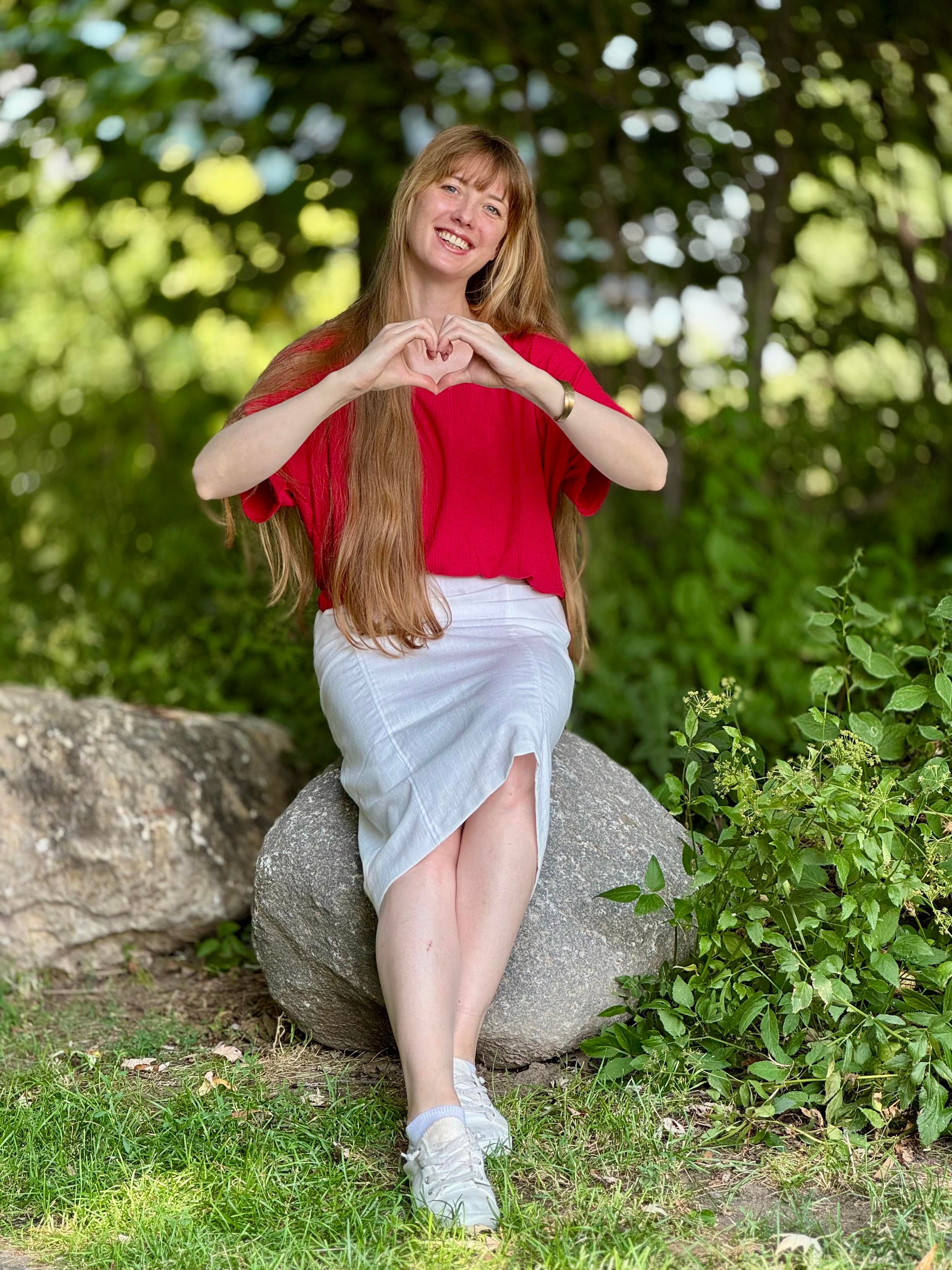 Picture of me in a red shirt and a white skirt, sitting on a rock, making a heart out of my hands and smiling at the camera. I am sitting in a park with a lot of greenery in the background, and I'm wearing my long blonde hair down.