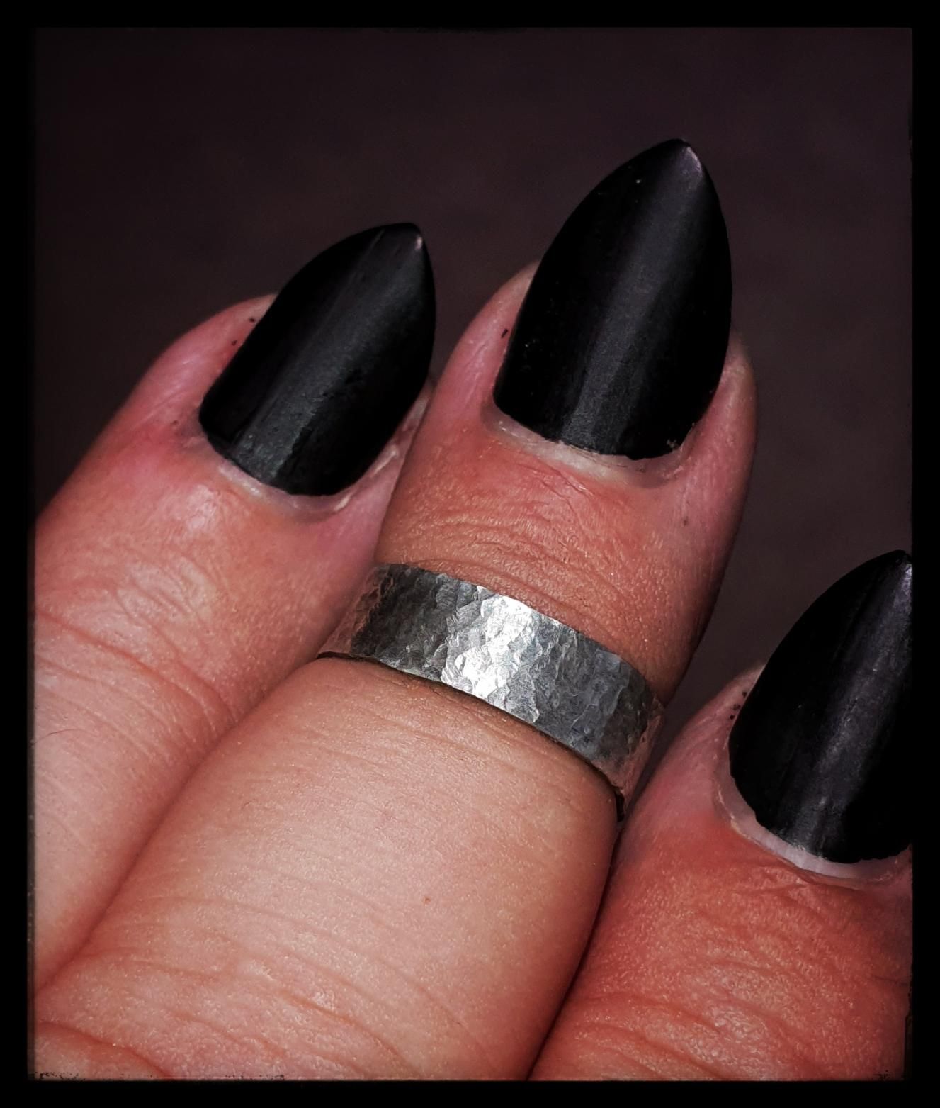 A close-up shot of three fingers adorned with long, matte black, stiletto-shaped nails. The middle finger wears a wide, hammered silver ring with a raw, rustic texture that catches the light. The contrast between the dark, elegant nails and the rough metallic sheen of the ring creates a bold, edgy aesthetic. The skin shows natural texture and subtle redness near the cuticles, adding a raw, unfiltered intimacy to the scene.