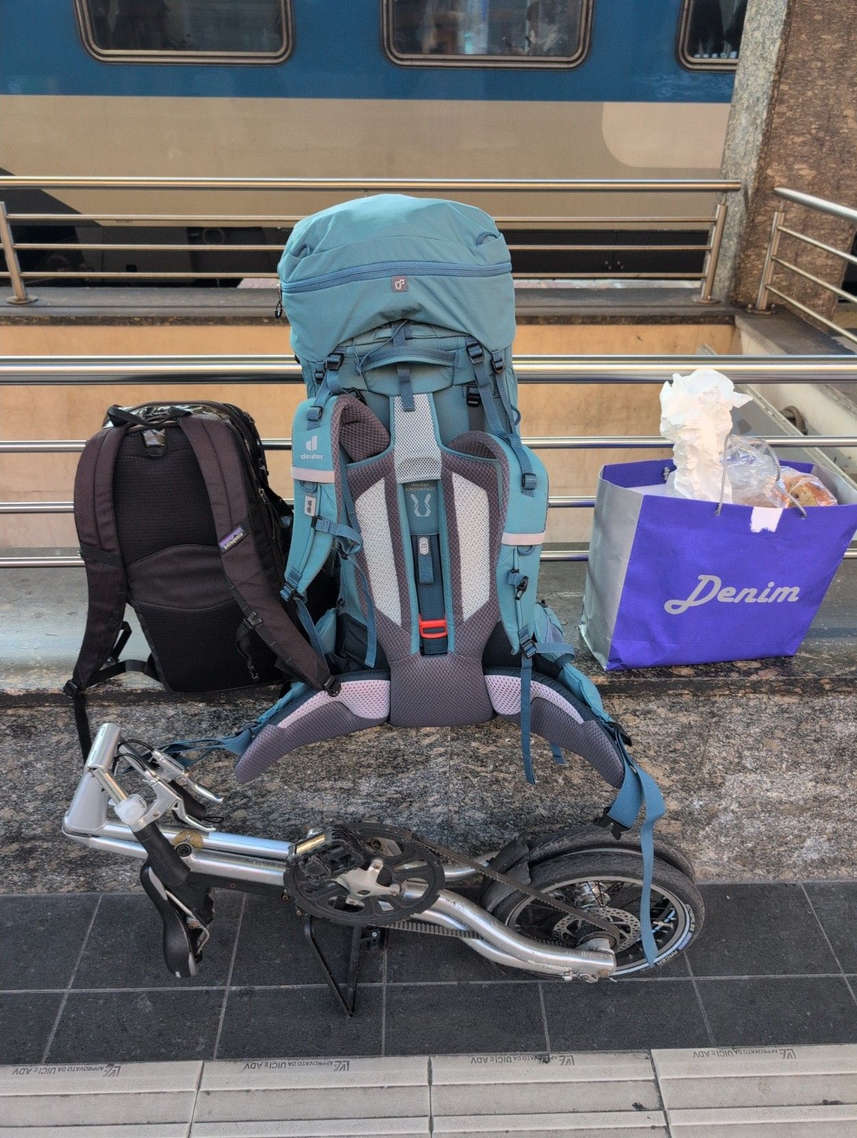 A photo of my 30L black backpack, my 65L light blue one, a bag of food cooked by my mum 💕 and my folding bike.