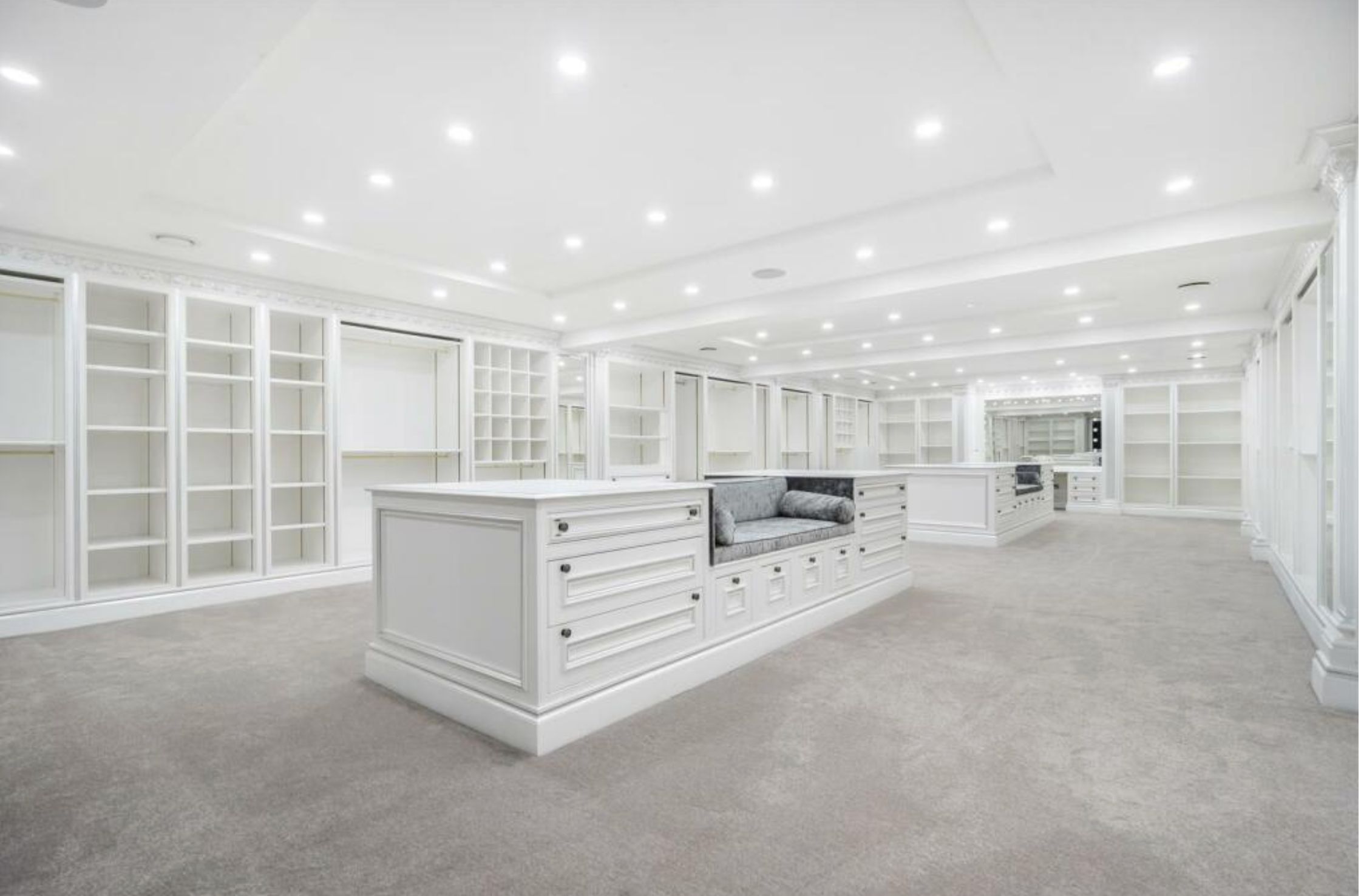 An absolutely gigantic closet with tan carpet and the walls surrounded by white shelves and rods for clothing, and islands in the middle with cushions for lounging and some drawers. There is a ton of empty space in it and there is no reason why there should be. 