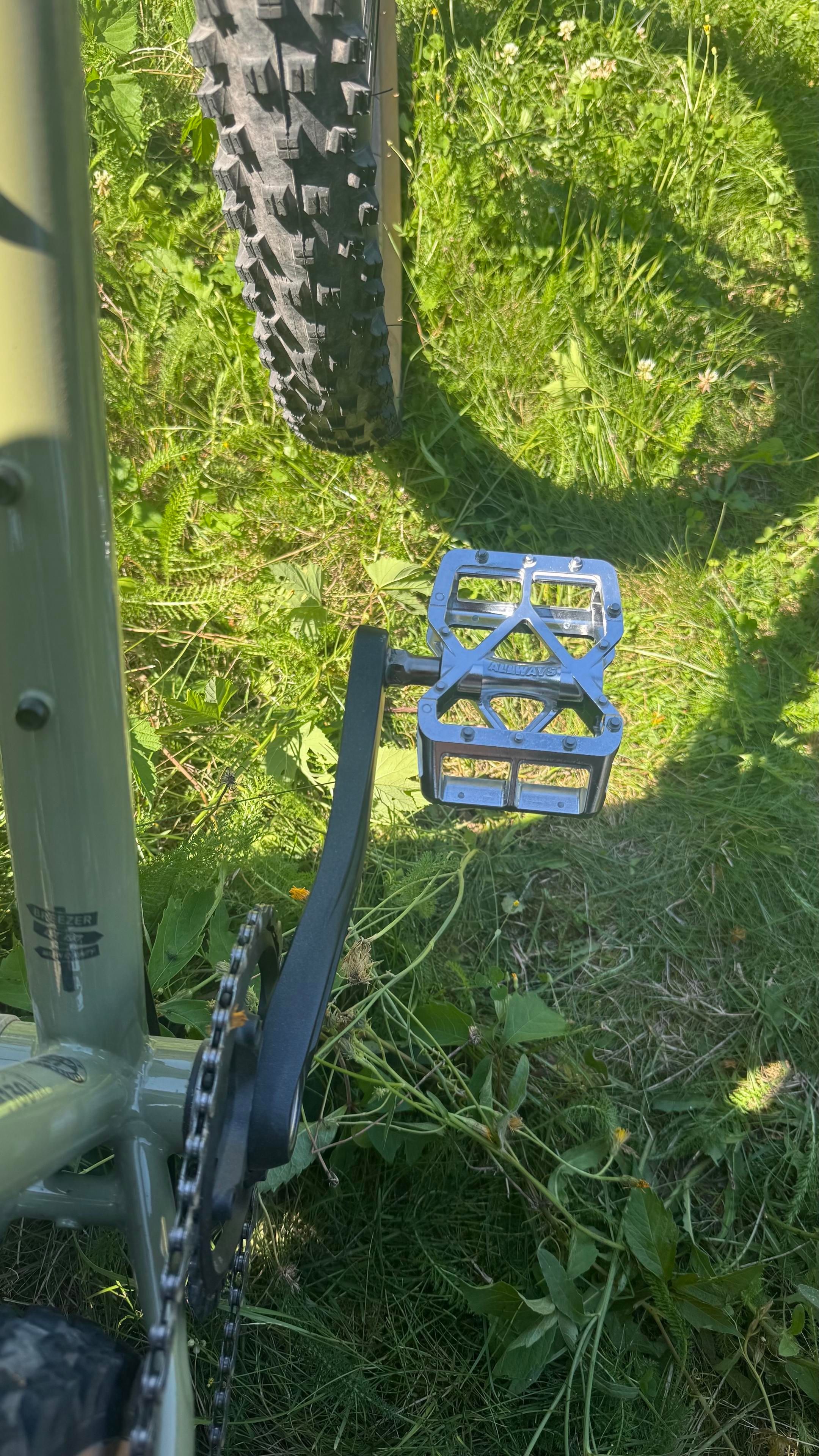 Nahaufnahme eines Fahrradpedals auf Gras, mit einer metallischen, strukturierten Oberfläche und einer sichtbaren Kette. Ein Teil eines Fahrradreifens ist ebenfalls sichtbar.