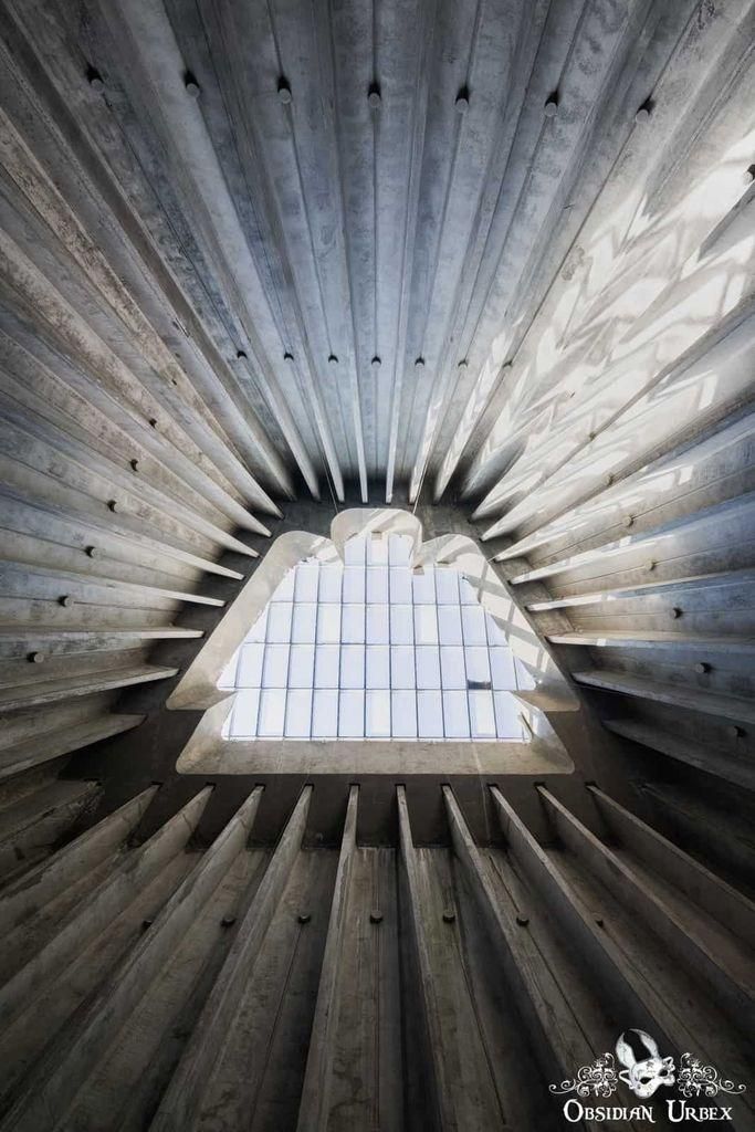 An upward shot reveals the dramatic triangular skylight of a Brutalist-style church, featuring ribbed concrete architecture.
