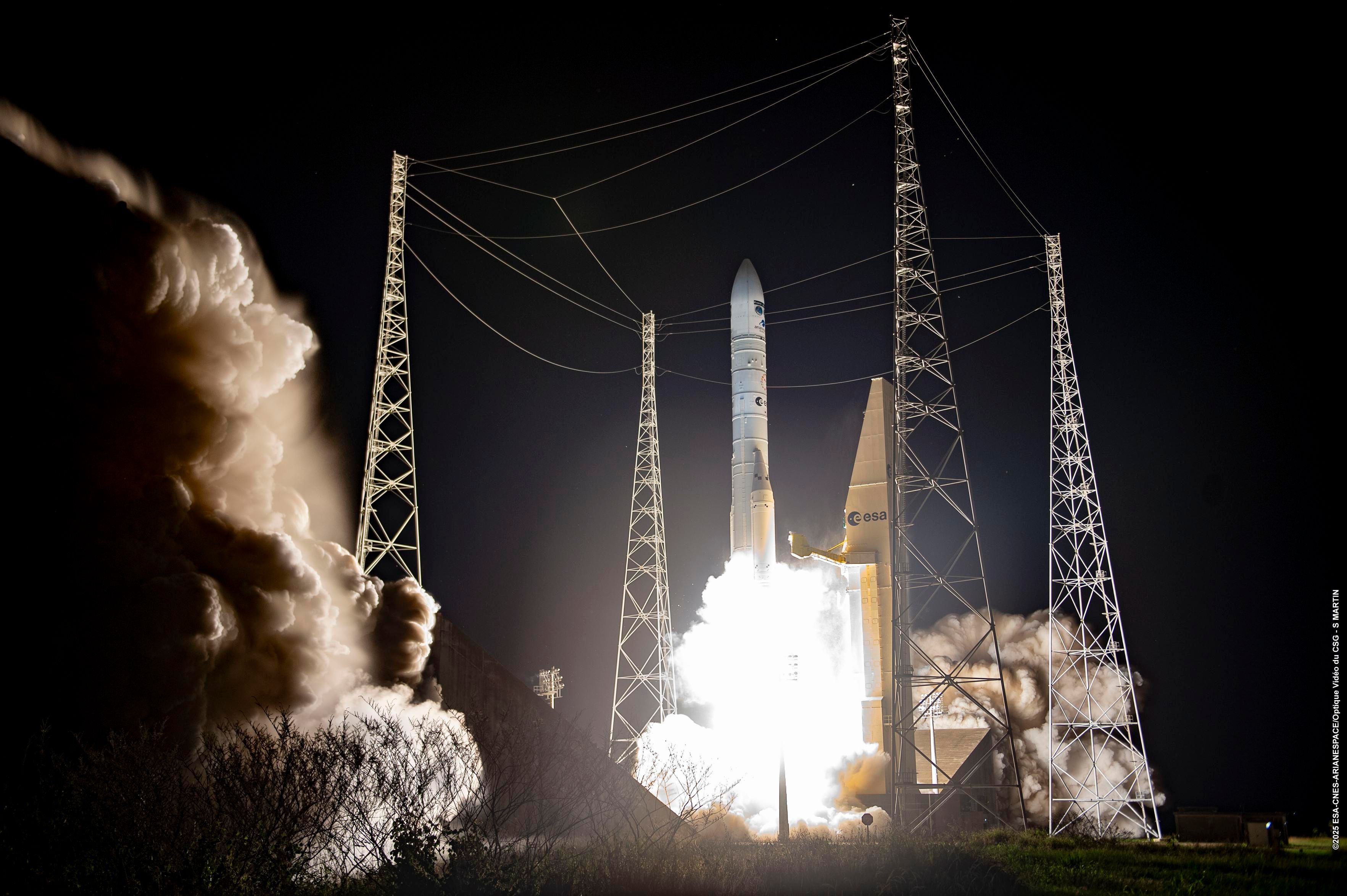 A Vega-C rocket lifts off at night from the European Space Agency’s launch site, surrounded by tall metal support towers and cables, with bright flames and thick clouds of smoke billowing into the dark sky.