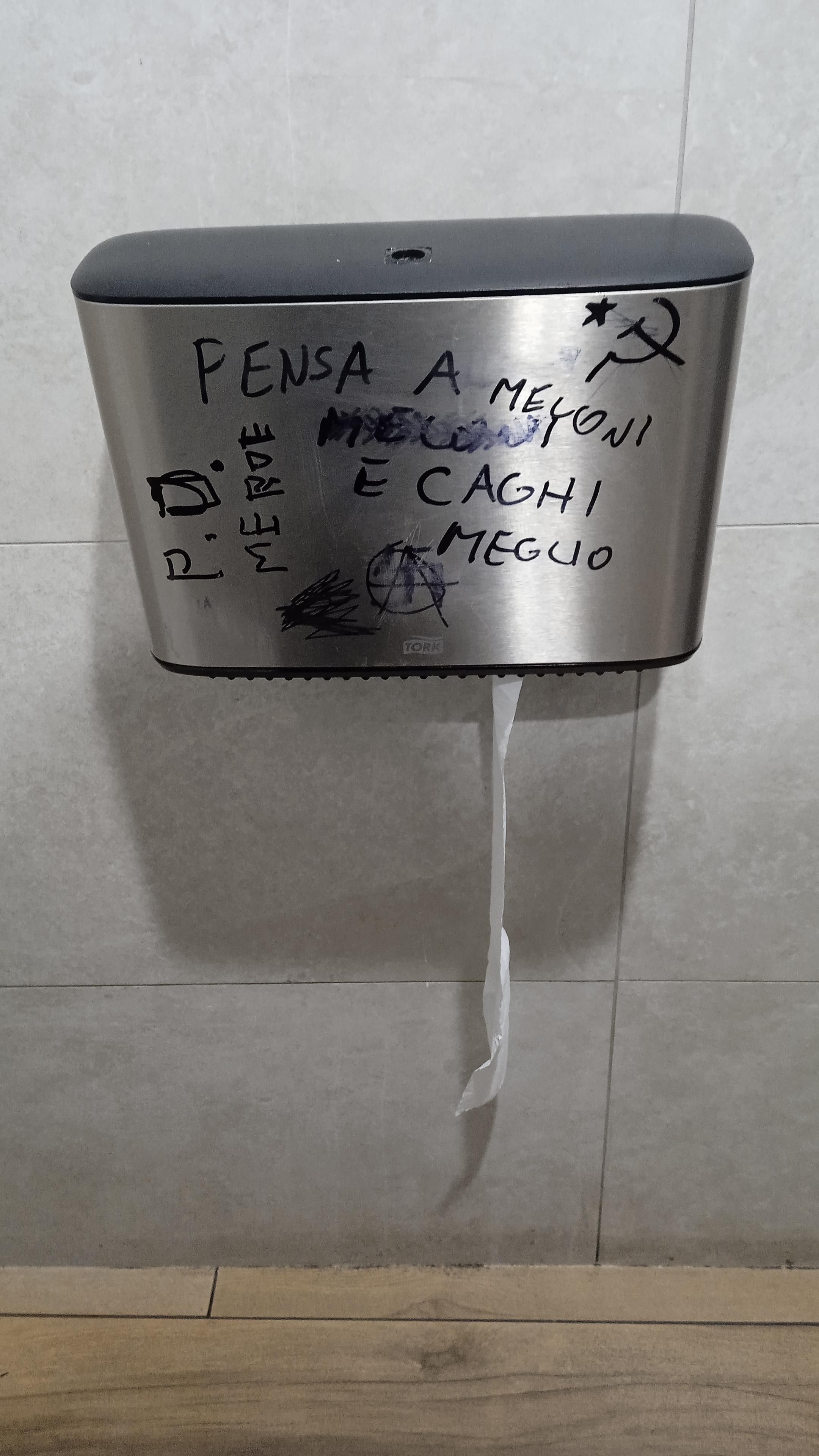 A metal toilet paper dispenser with a marker pen writing on it that says "pensa a meloni e caghi meglio" . In the upper right corner a hammer and sickle with a star. On the left a writing "pd merde"