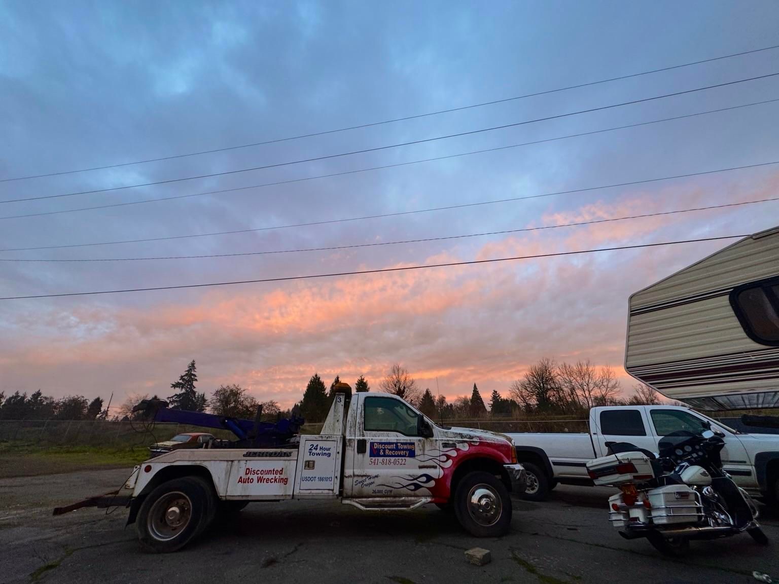 An old tow truck with a colorful design is parked beside a white pickup truck and a motorcycle. The background features a cloudy sky with pink and blue hues at sunrise, along with some trees and power lines.