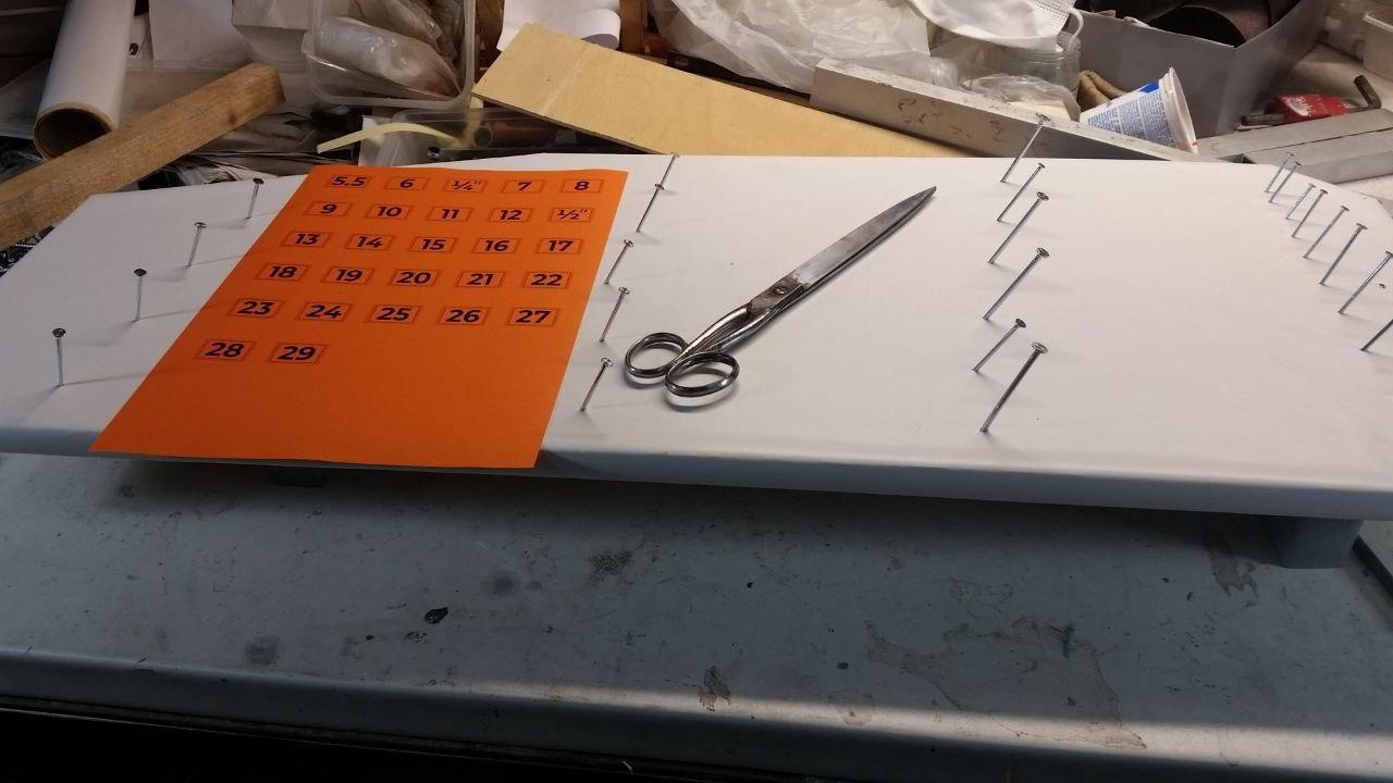 Pannello per appendere le chiavi appoggiato al tavolo, con tutti i chiodi conficcati.
Verso sinistra un foglio arancione con tutte le etichettine da ritagliare e fissare con lo Scotch Magic (ma prima bisogna dargli una leccata di colla stick sotto o lo scotch le "attira" e le madonne volano), a fianco un paio di forbici per tappezziere (lunghe 26 cm, prima che chiediate).