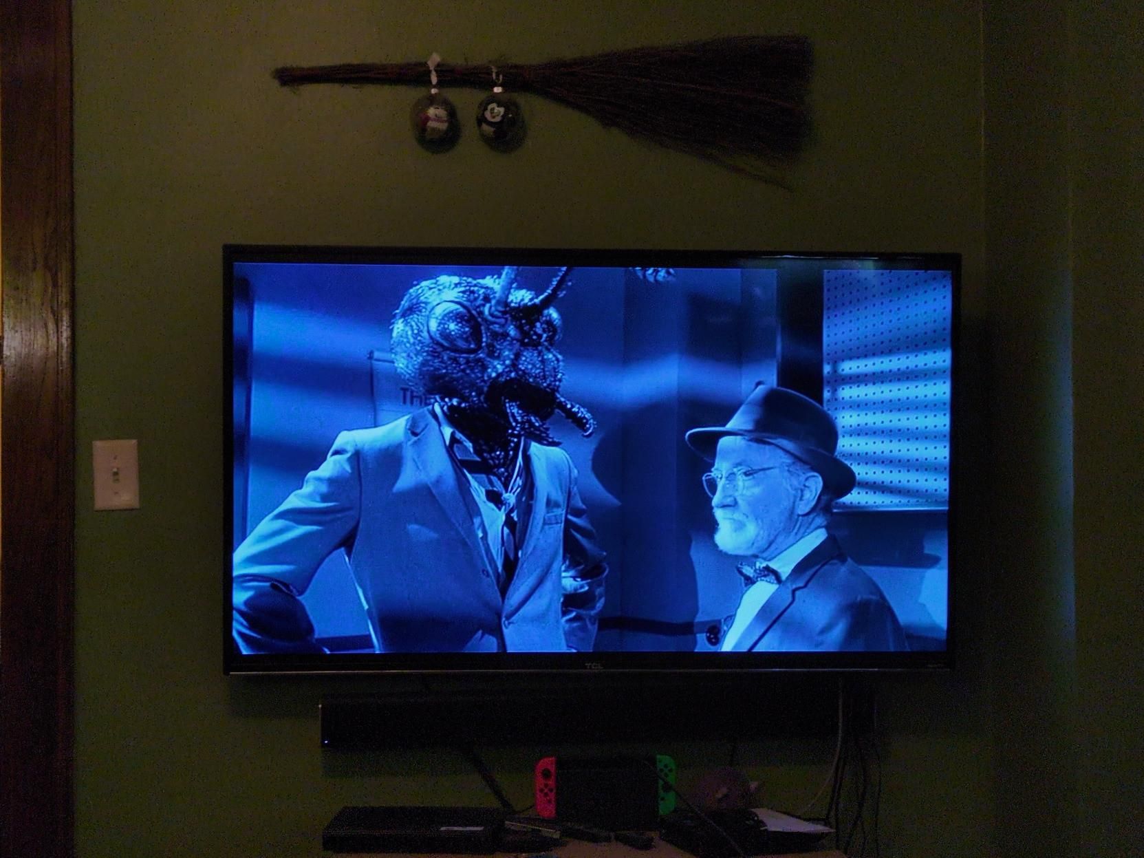 A photo of a television set on wall. On screen, a man in a suit with a large ant head, looks indignant at a disinterested older man in a suit. 