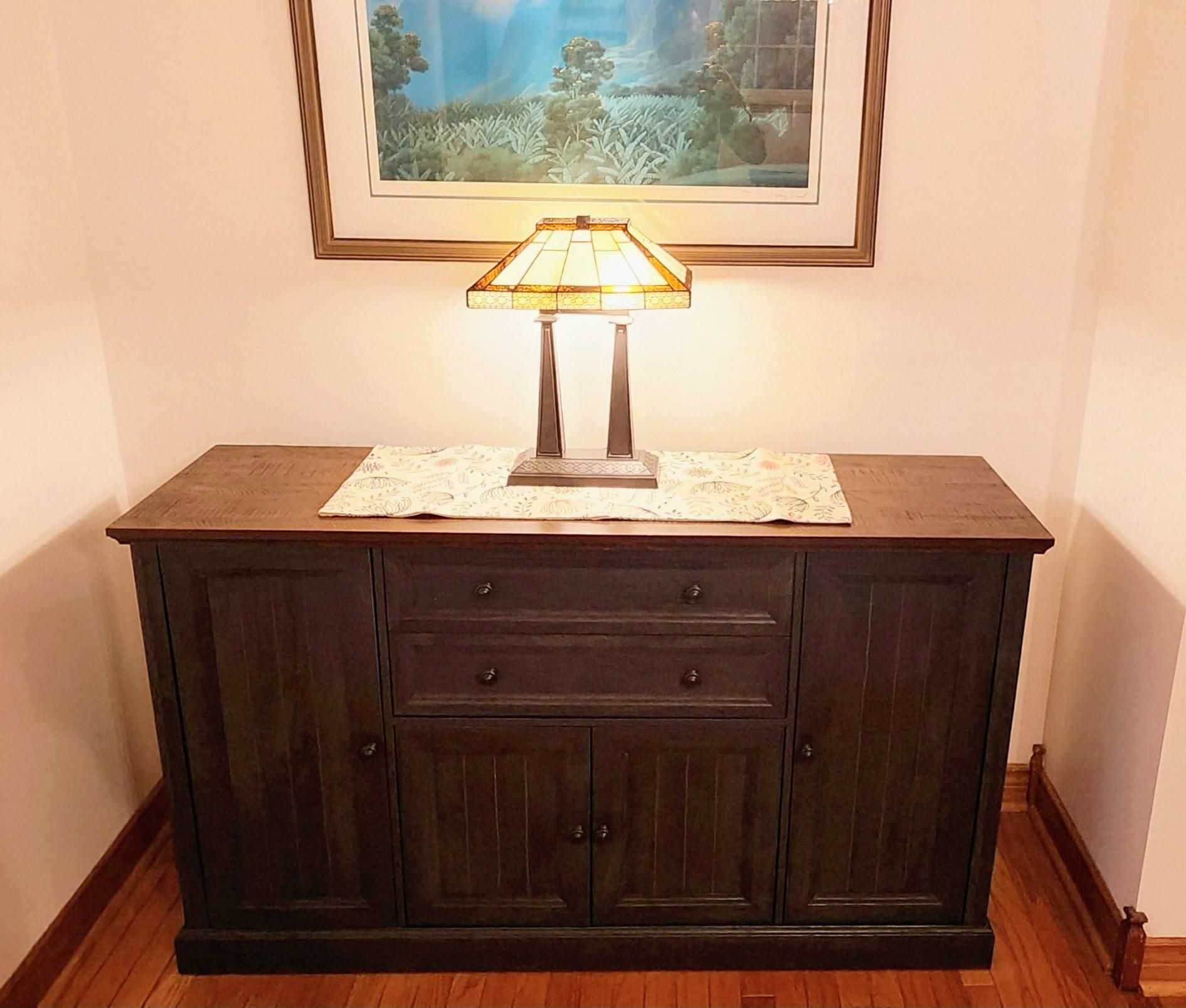 A picture of the sideboard now completed. It is just under 2 metres long and less than a metre tall. There are two large cupboard doors, the height of the sideboard, on the outer sections. The middle section has two wide drawers with two smaller cupboard doors underneath.  The color is a dark brown/black wood grain stain while the top mimicks a dark barnwood finish. A replica Tiffany lamp sits on top, turned on, and a print of a painting hangs above.