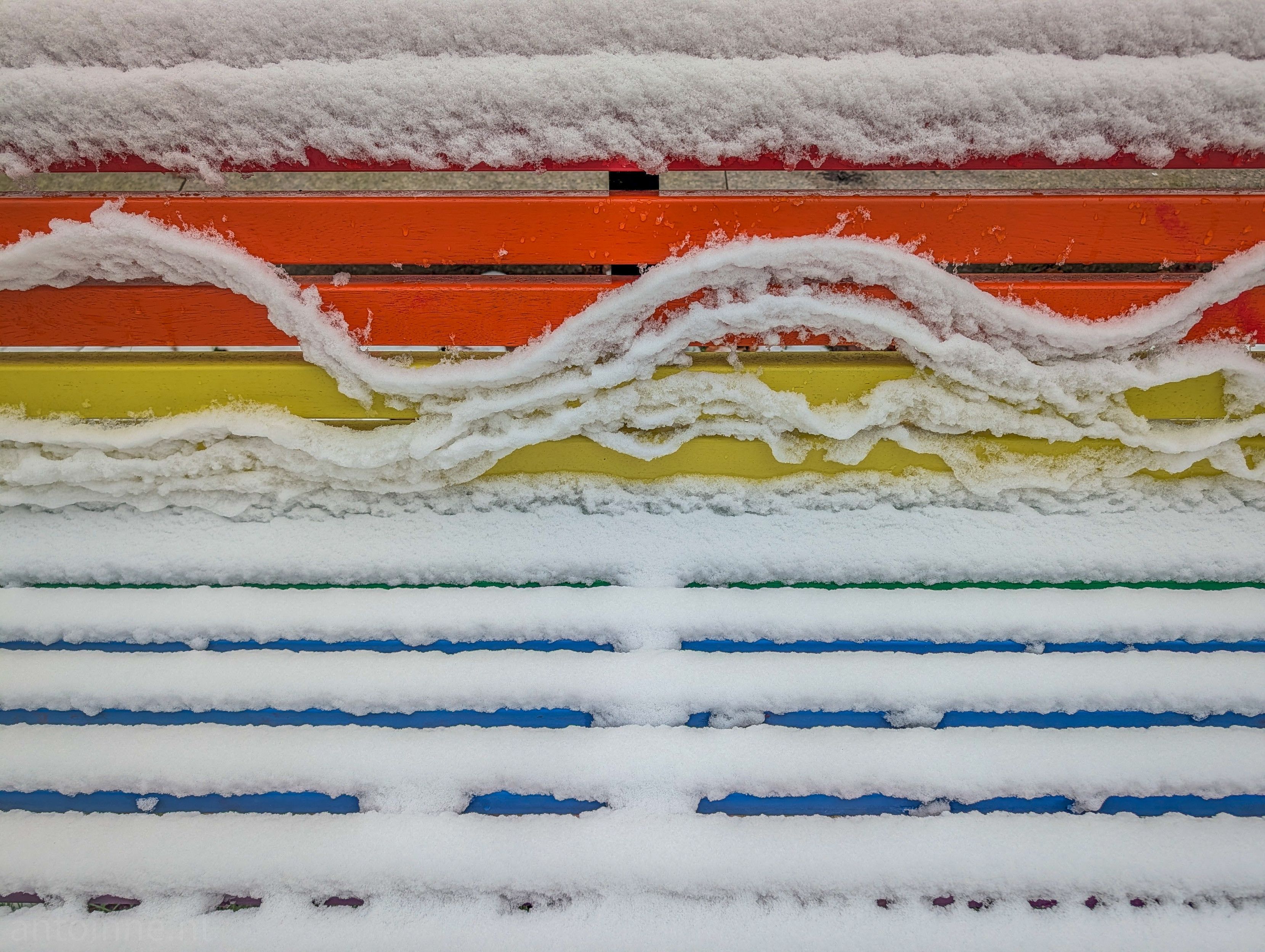 A vibrant, multi-colored wooden bench partially covered in fresh snow. The bench features horizontal slats painted in the colors of the rainbow, creating a striking contrast against the white snow.

On the upper slats, the snow has begun to slide, forming elegant, wave-like curves and "ribbons" rather than a solid blanket.

A rainbow bench is a bench in a public space that is painted in the colors of the rainbow flag of the LGBT+ movement. Its function is similar to that of a rainbow crosswalk.