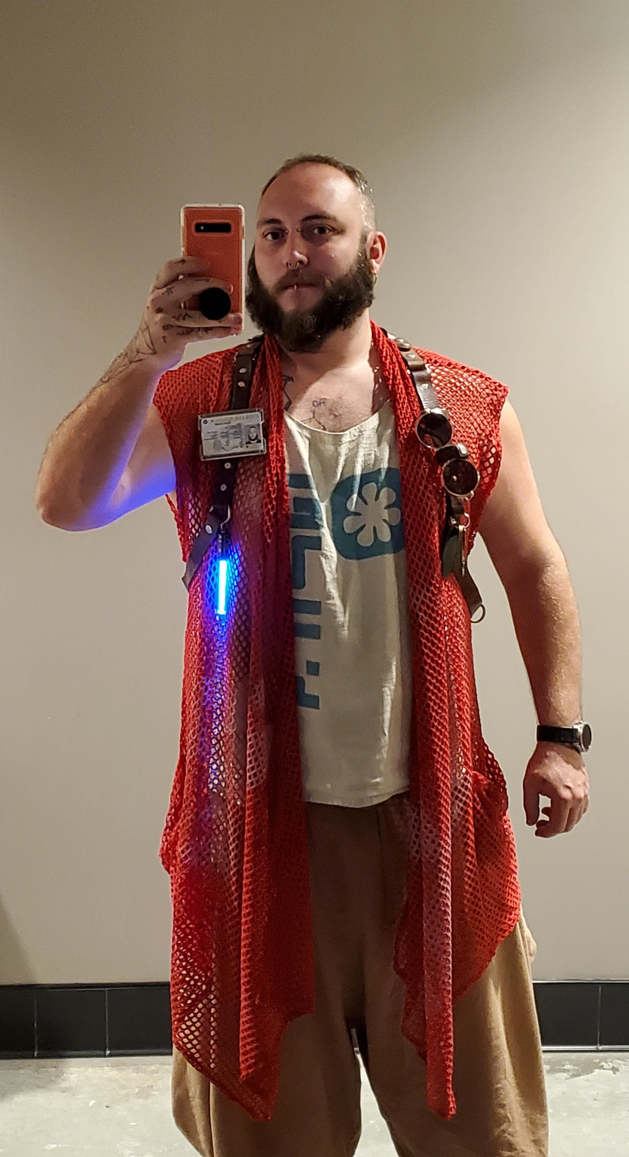 Selfie. Wearing a long red fishnet vest with white logo print, brown harem pants, and a white linen sleeveless.