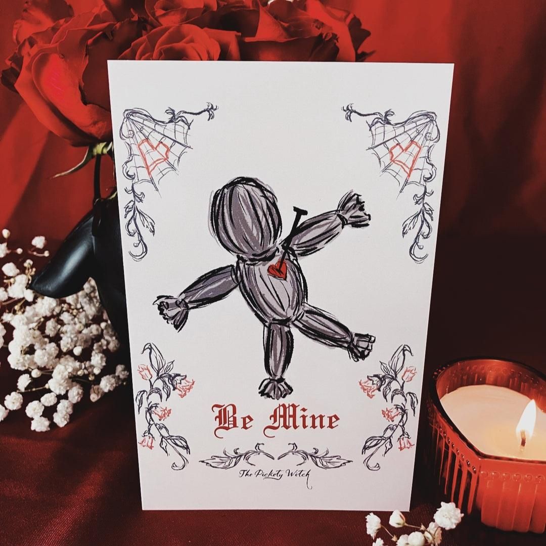 An illustrative Valentine's Day card with a gothic aesthetic featuring a hand-drawn voodoo doll with a pin through a red heart on its chest. The card displays the text "Be Mine" in red gothic calligraphy and is set against a romantic backdrop of red roses, white baby's breath, and a lit candle.