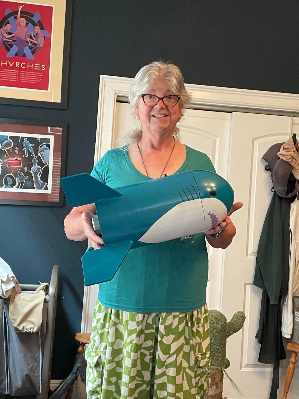 A smiling white woman with grey hair and glasses holding up a stubby, four fin model rocket decorated like a Blåhaj