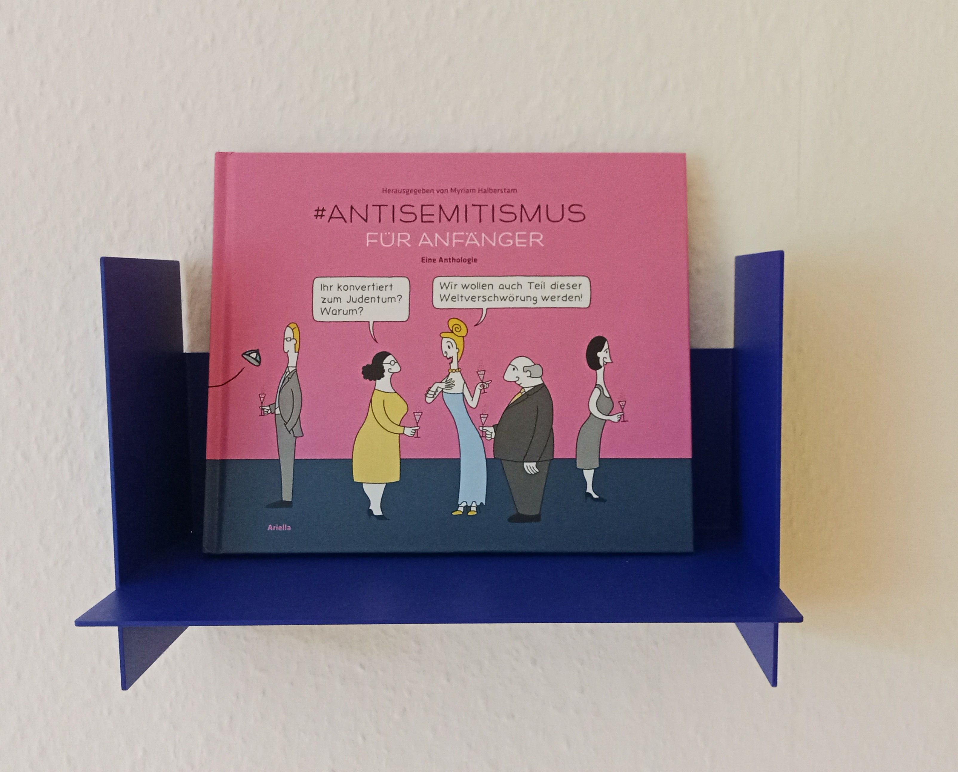 in einem kleinen yves klein blauen metallregal von tiptoe steht wie in einem bilderrahmen das quietschlila karikaturenbuch "antisemitismus für anfänger". auf dem cover die zeichnung reicher personen auf einer party im smalltalk. "Ihr konvertiert zum Judemtum. Warum?" "Wir wollen auch Teil dieser Weltverschwörung werden."