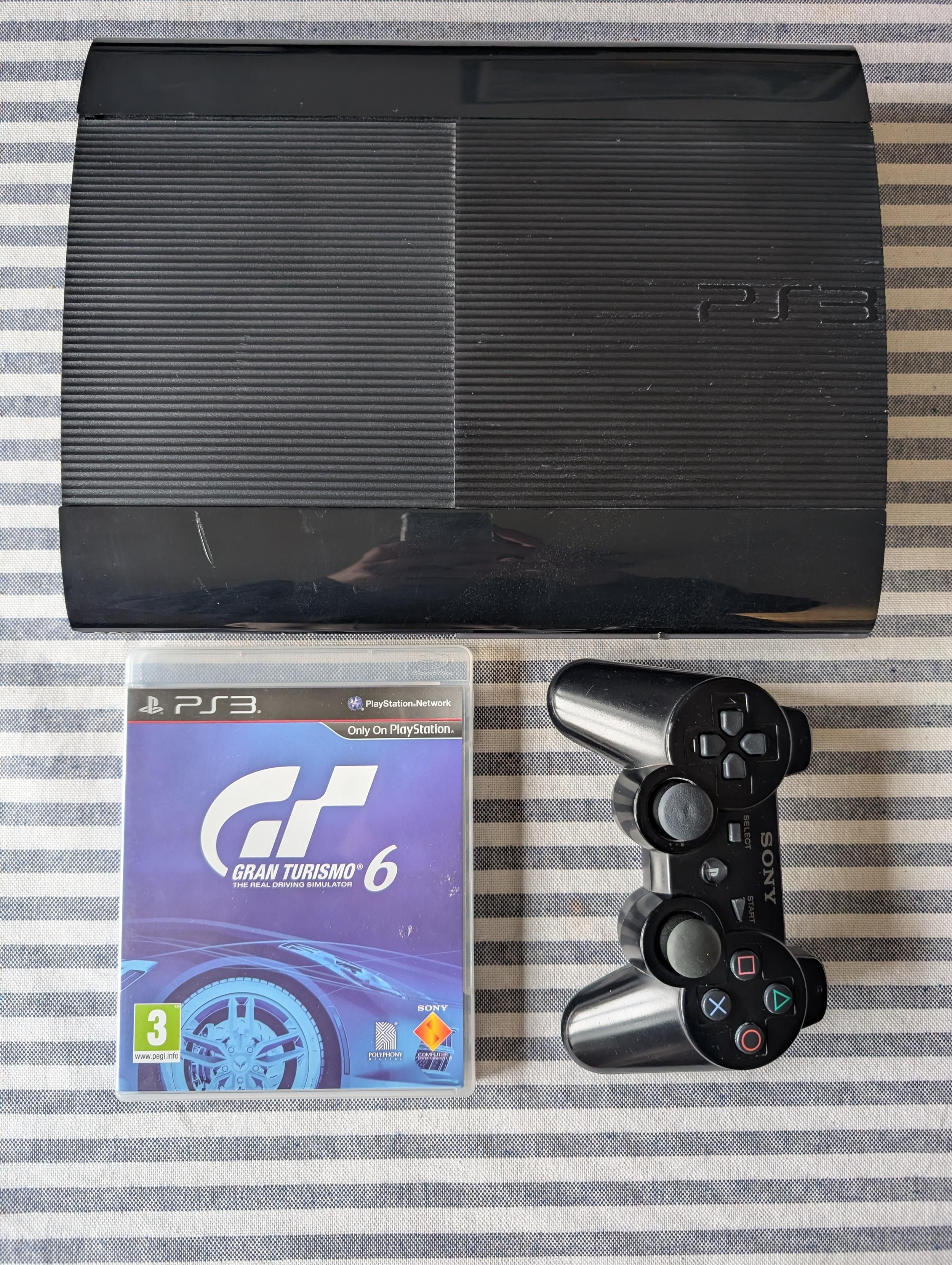 Fotografia di tre oggetti disposti su di un tavolo: una console nera, un gioco con copertina blu e un gamepad nero.