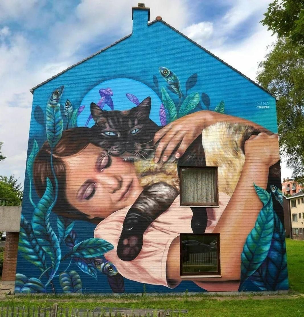 A brightly coloured house end painted with a mural of a woman holding her cat warmly, eyes closed, while laying down. The background is turquoise with plants around her, giving a warm yet ethereal experience