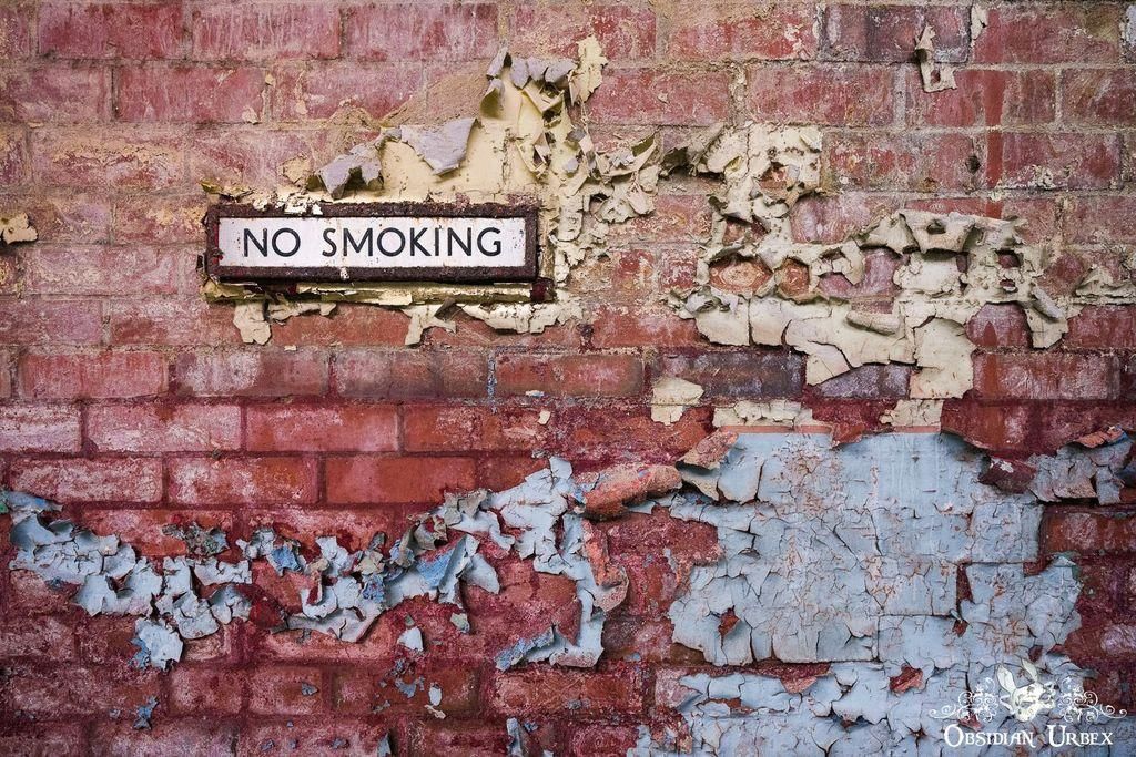 A grungy brick wall with peeling paint surrounds a "NO SMOKING" sign. The paint is flaking off in layers, revealing the brick underneath.