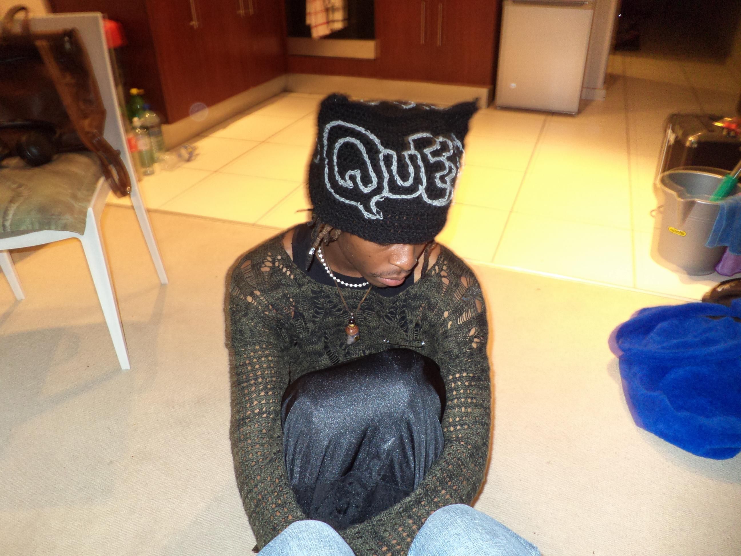 a cute photo of me sitting on the floor with a green knit top and black skirt, with a cute crochet hat with the words "queer" 