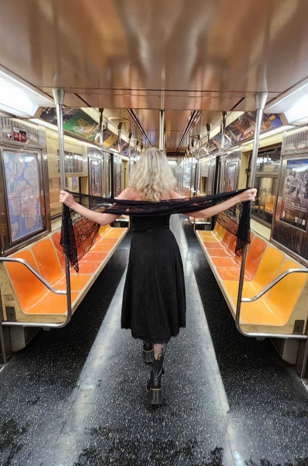 Me from behind standing in a clean empty orange subway car. My arms are outstretched holding the poles with a black shawl. I am wearing a strapless calf length black dress and boots with a block here. My hair is down, mid back length and white. 