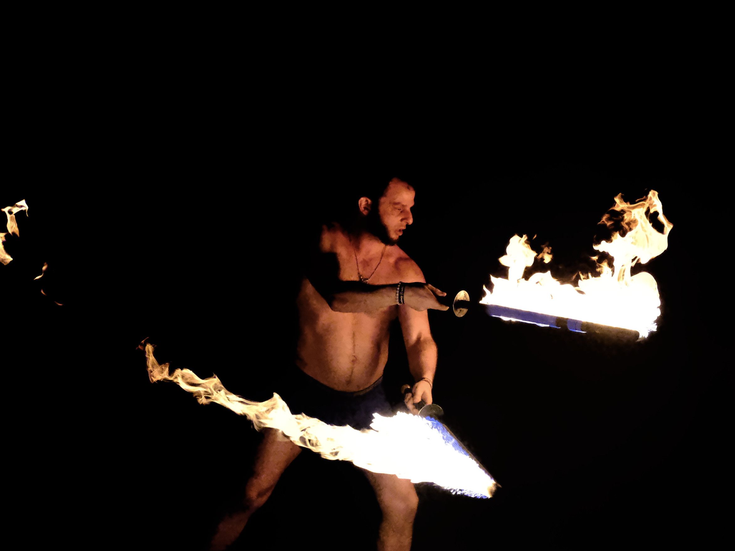 A man with an angular face and prominent, bold nose, wearing only shorts, wields two flaming swords, each pointing in a different direction. His red-orange skin (reflecting the flames) stands out against a pitch-black background.