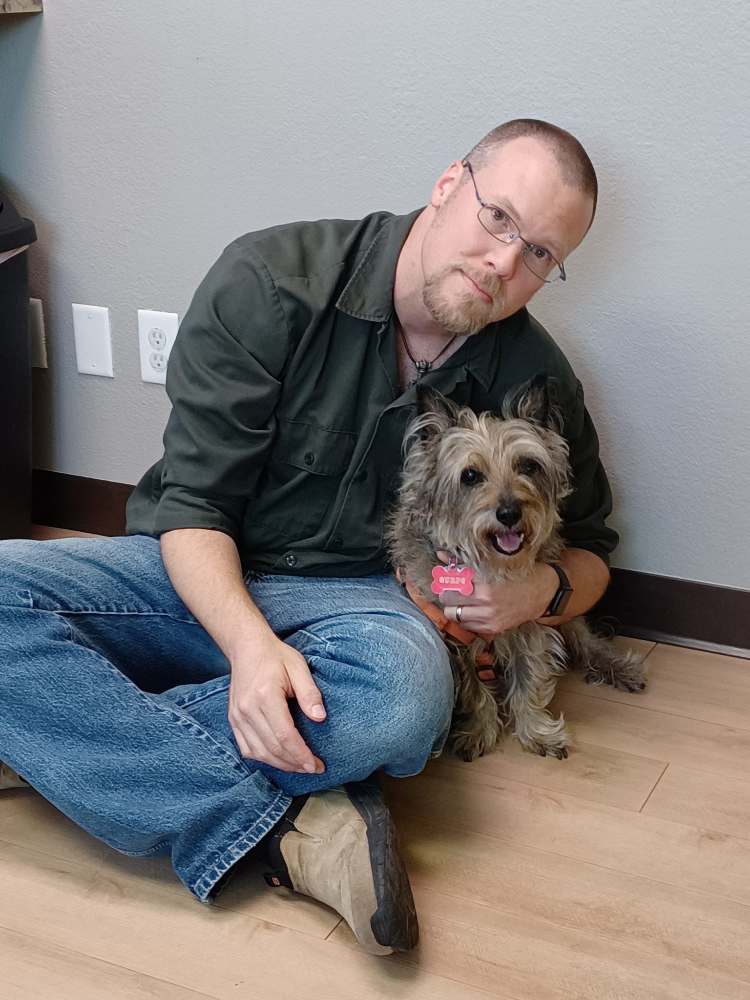 Photograph of a middle-aged man with short hair and glasses, wearing a green work shirt and jeans hugging a brindled (brown and tan fur) cairn terrier in the vet's office.