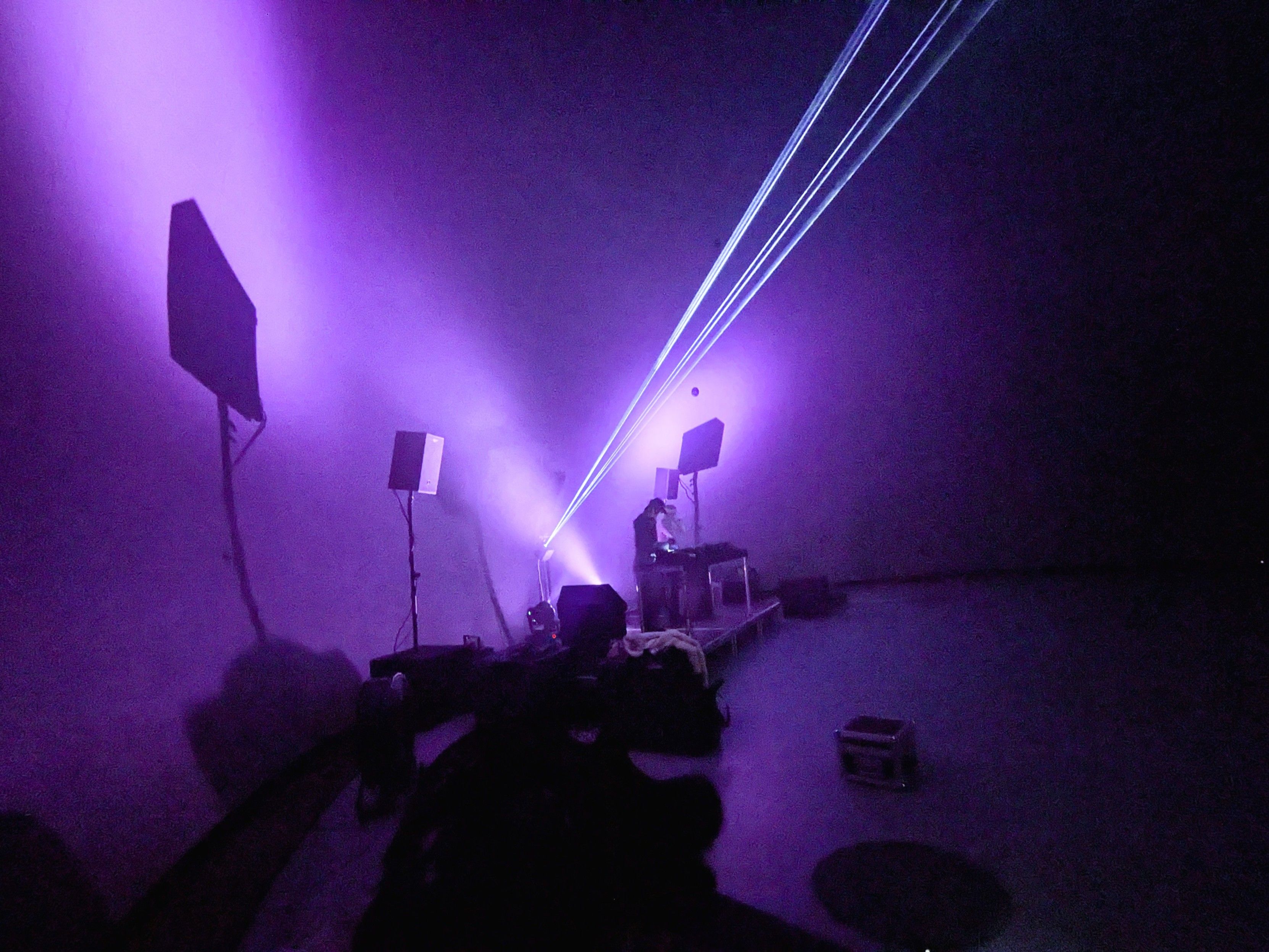 Wide angle photo of a large dome shaped venue with a stage, moving heads, haze and lasers. The lights are pink and someone is setting up gear on the main table on the stage