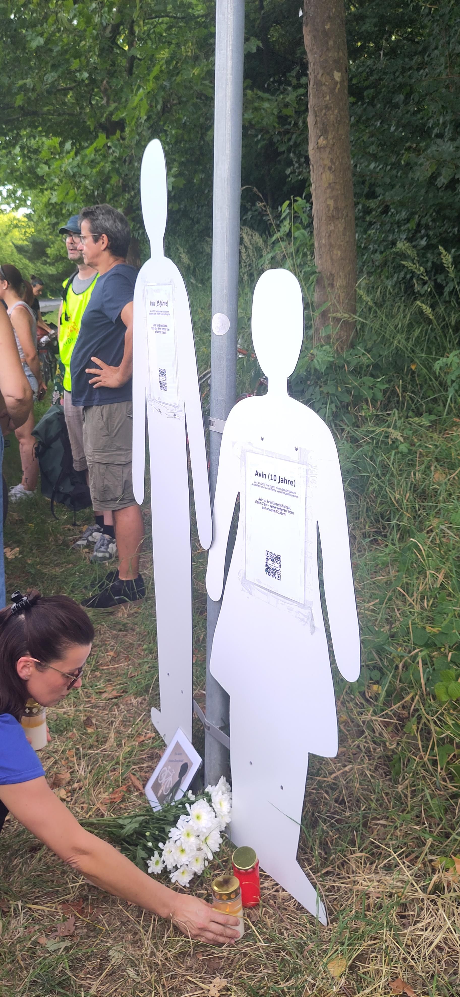 Zwei etwa lebensgroße Symbole für Avin (10 J.) und Luis (25 J.) Zu Füßen liegen Blumen und eine Karte. Eine Frau stellt eine Grabkerze auf.