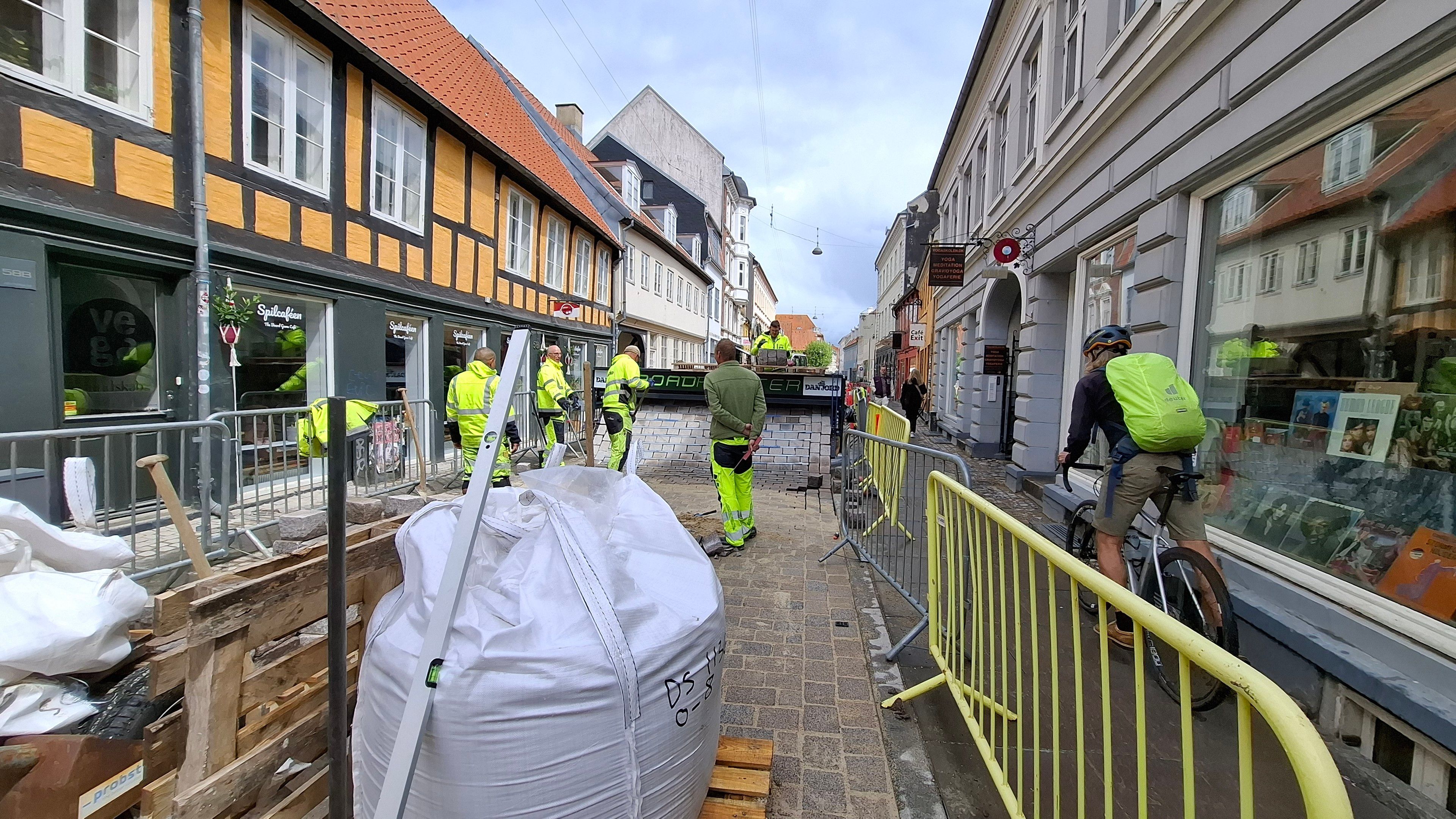 Seen from the back, behind building paraphernalia and a huge bag of concrete or some such - a vew of the brick road being wound out of the machine. A cyclist is just passing on the pavement alongside. Same general description from first photo now follows, so skip if already heard: Vestergade, mid-city Aarhus, Denmark. A lovely narrow old street that is, after a few years' trial, being turned permanently into a Quiet Street - cars may drive here, but only slowly, and bikes and pedestrians have priority. The council is planting trees, widening the pavement café and seating spaces, and laying a nice new chessboard-like brick surface. The surface is designed to slow down bikes too.  It is the first place in Denmark that is being paved with a Dan Jord RoadPrinter - a Dutch brick paver that rolls out complete stretches of road from a machine, as if you are winding a continuous sheet of paper out from a huge roll of paper. Photo taken with permission - I have turned into one of those Auld Lads who stand there, hands behind my back, watching them at work, fascinated by the process, interrupting them with questions including 'may I take some photos?