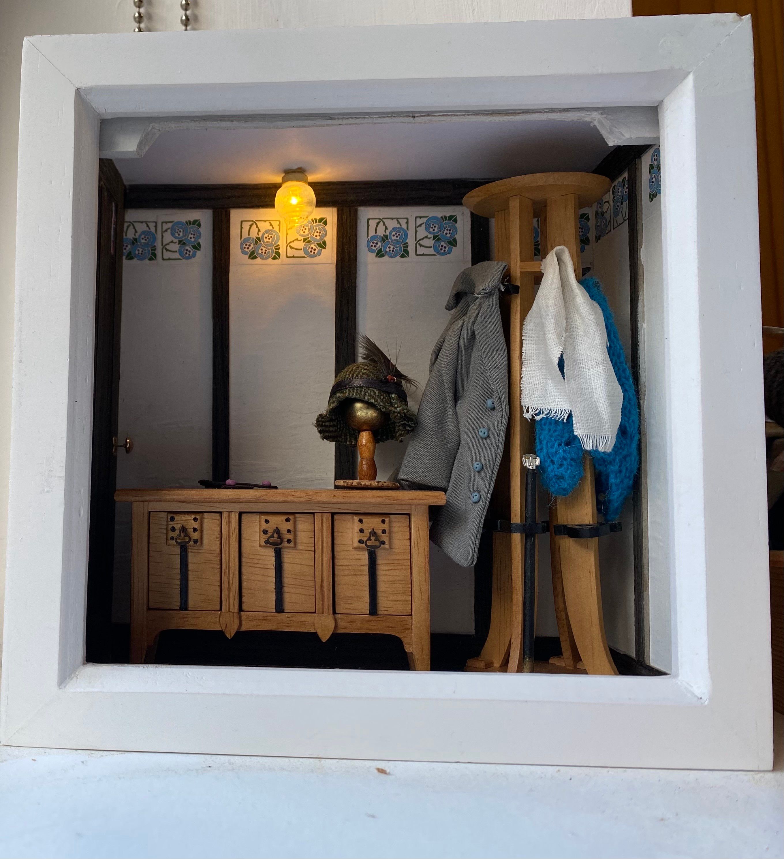A miniature scene depicting an entrance hallway with a wooden bench, a hat rack with a coat and a scarf, and a light fixture. The walls are decorated with floral patterns in the Glasgow style.