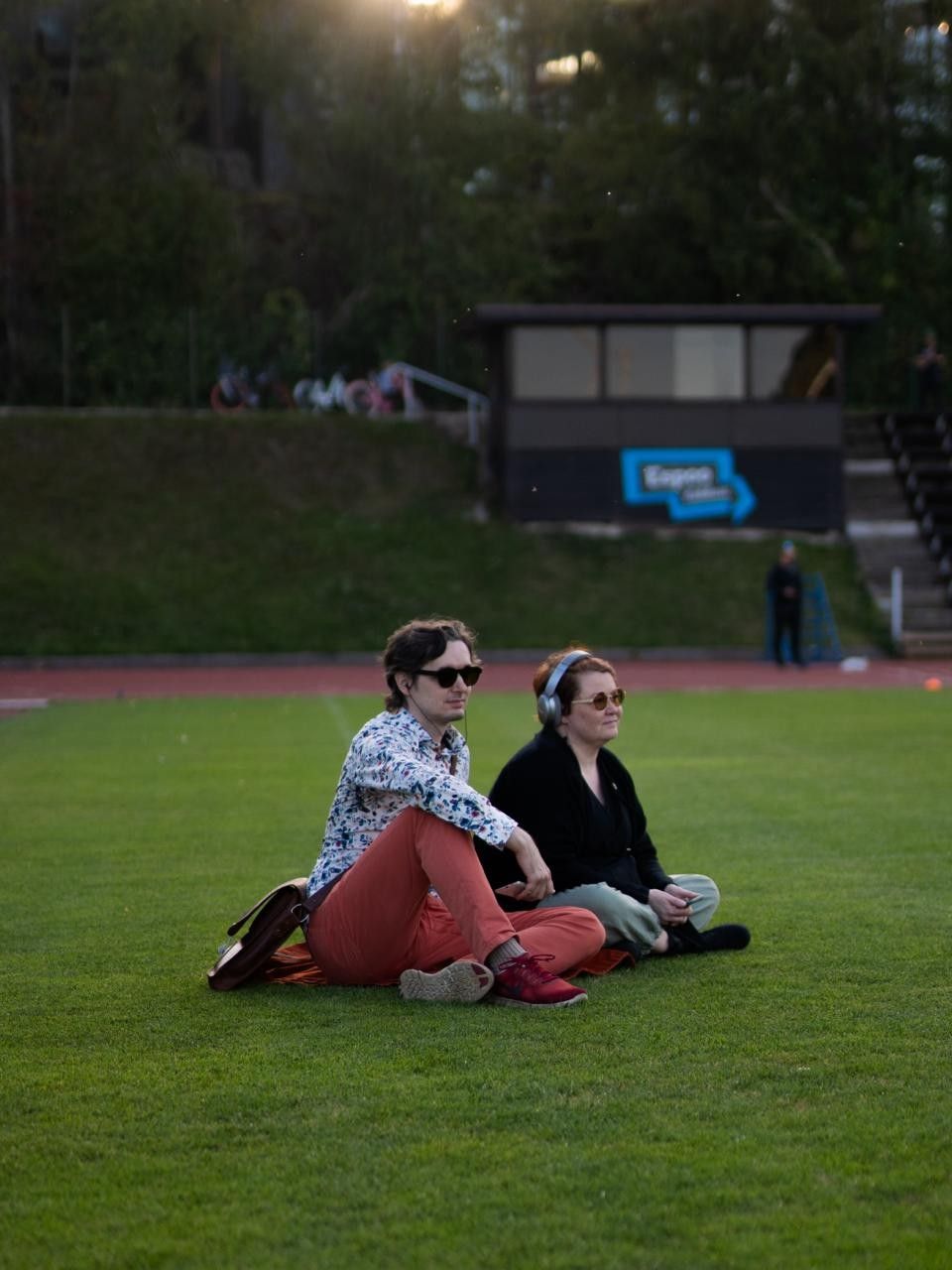 Kuva kahdesta henkilöstä istumassa urheilukentän viheriöllä kuulokkeet päässä ja aurinkolasit silmillä.

Etualalla minä värikkäässä kauluspaidassa ja punaisissa housuissa nahkainen olkalaukku mukana.