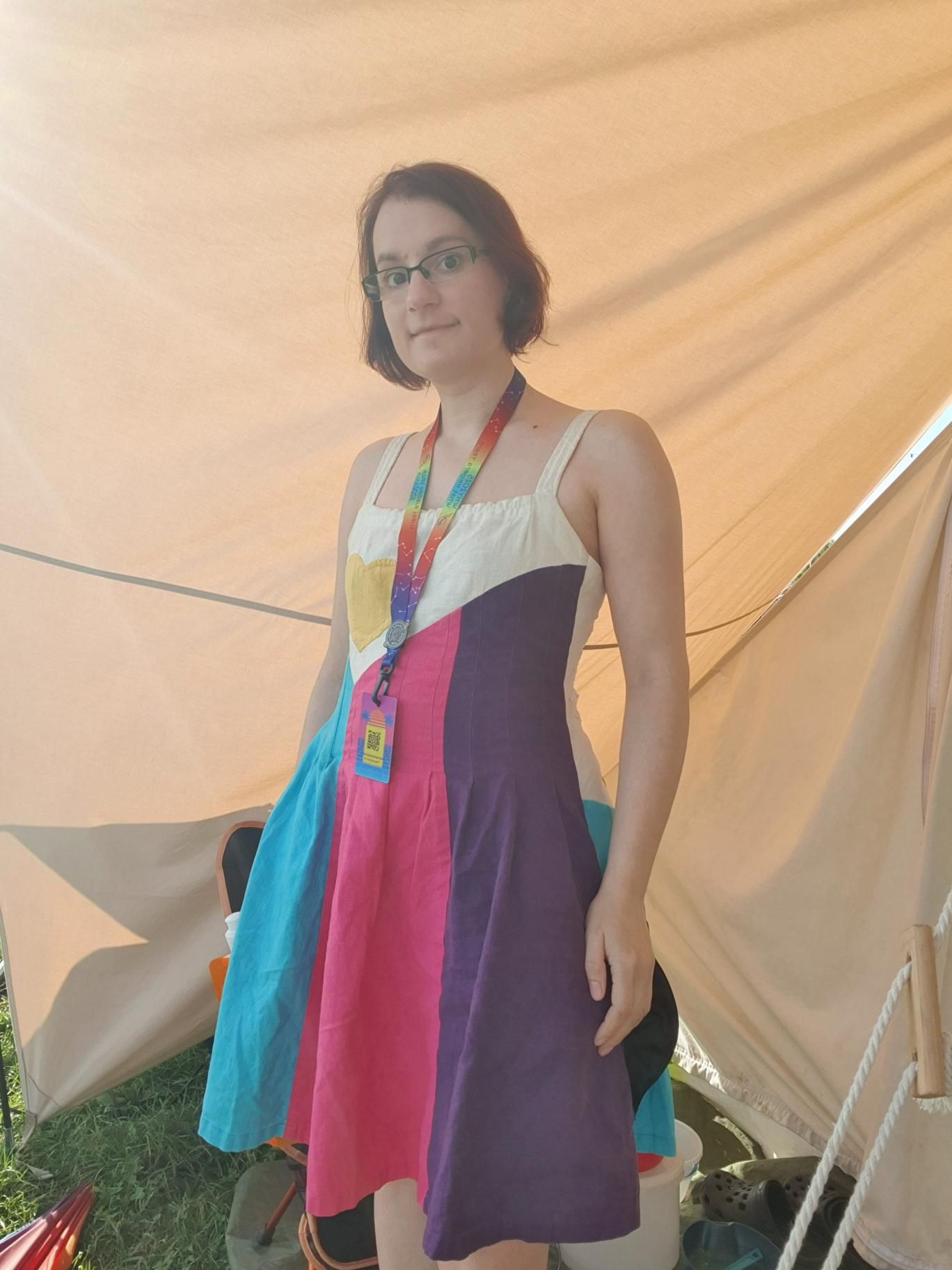 I'm standing under the canopy in front of a tent, wearing a knee-length sundress made of linen. It has cyan, pink and dark purple stripes in the waist and skirt part, and a white triangular shape with a yellow heart on it in the bust part. The shoulder straps are white as well. A colorful lanyard with a plastic card (angel badge) is hanging around my neck.