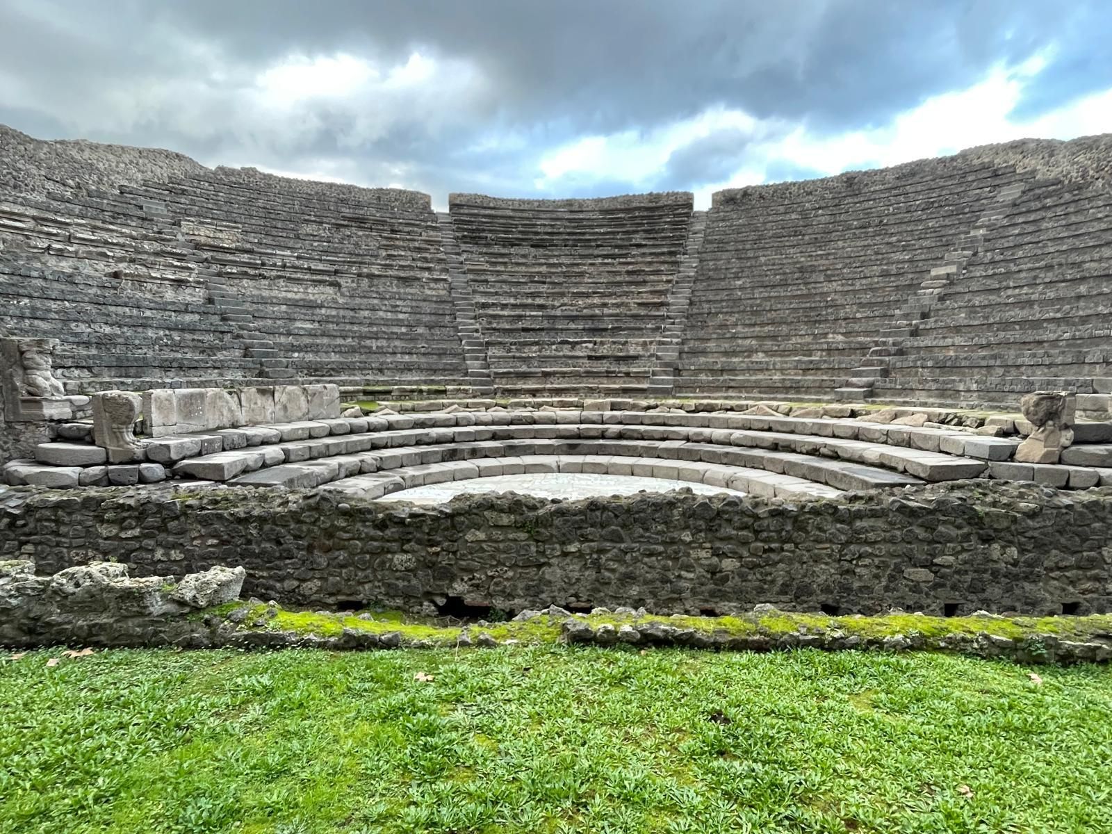 Vista generale del teatro piccolo. In primo piano una striscia di prato verde. Al centro la scena e le scalinate, divise in cinque settori dalle salite. Tutto molto ben conservato. In alto si intravede il cielo coperto di nuvoloni grigi
